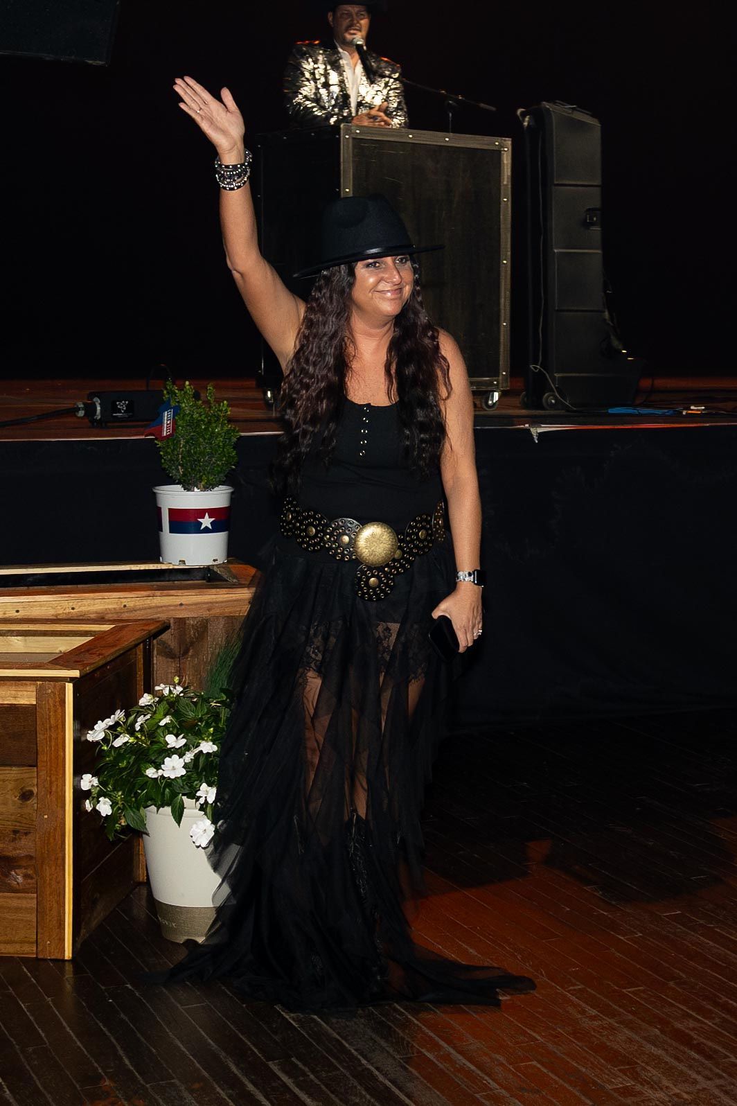 Woman in black dress waves from stage, DJ in background.