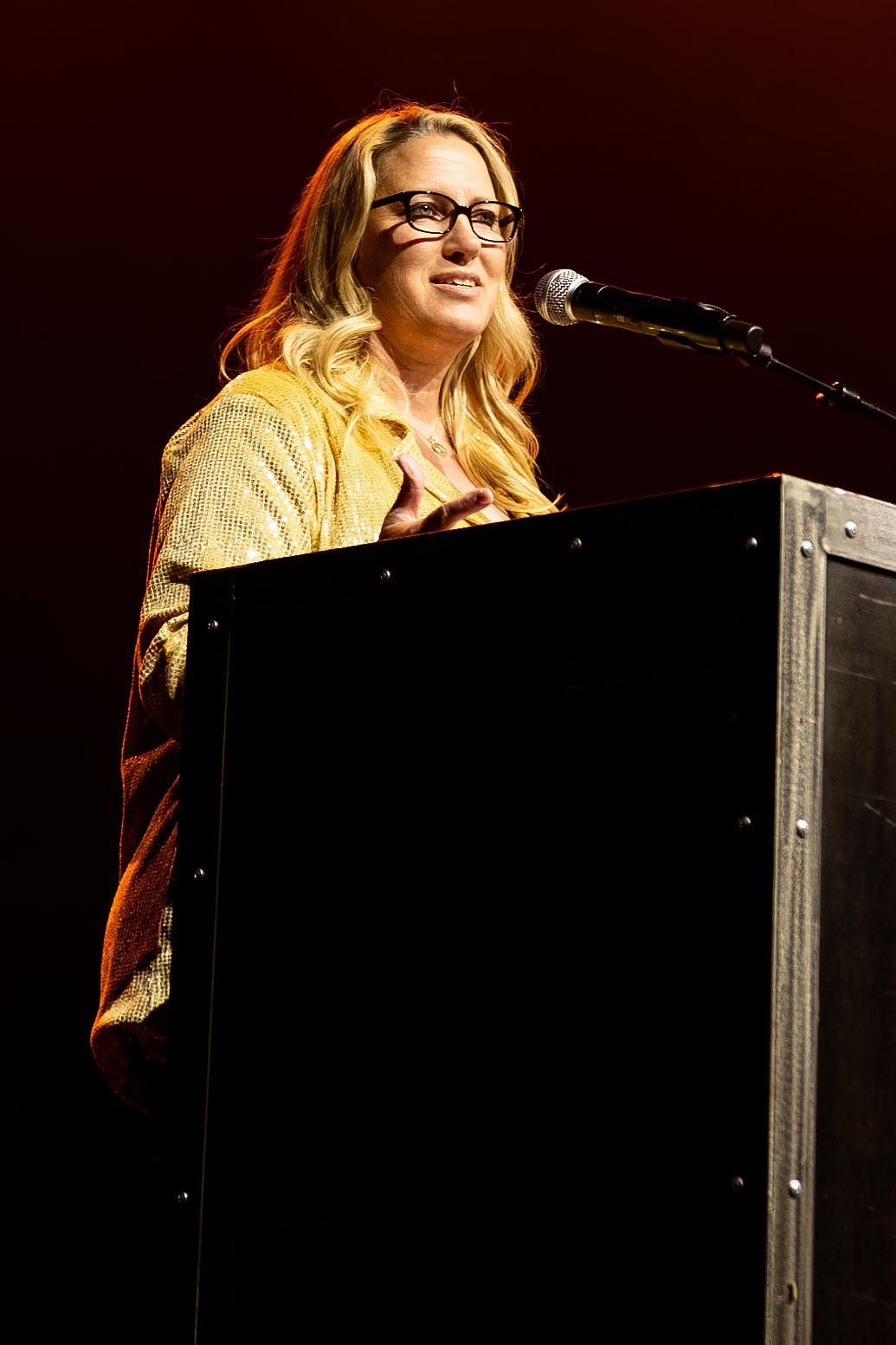 Woman with blonde hair and glasses speaking at a podium on stage, wearing a yellow sweater.