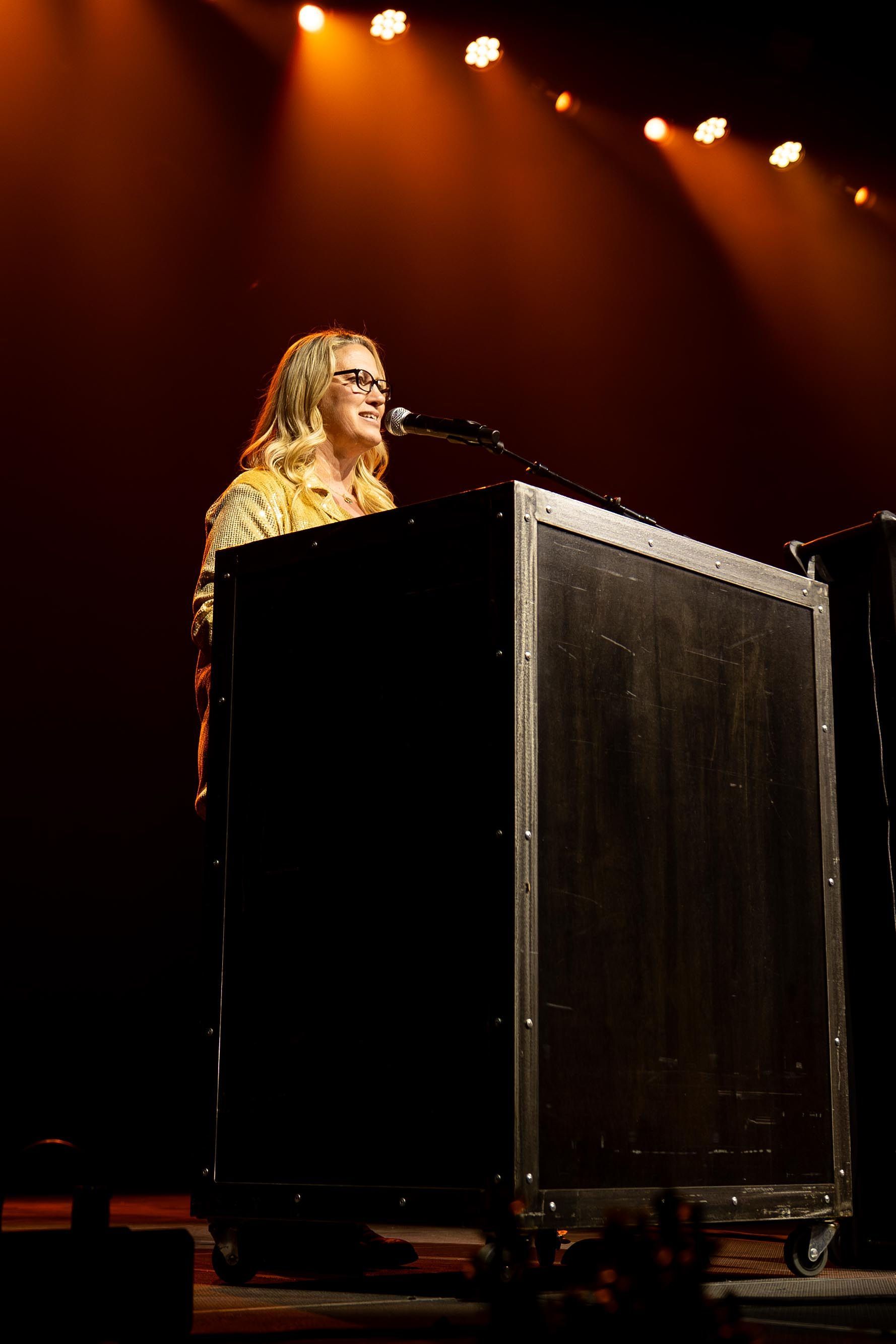 Woman with blonde hair and glasses speaking at a podium on a stage with orange lighting.