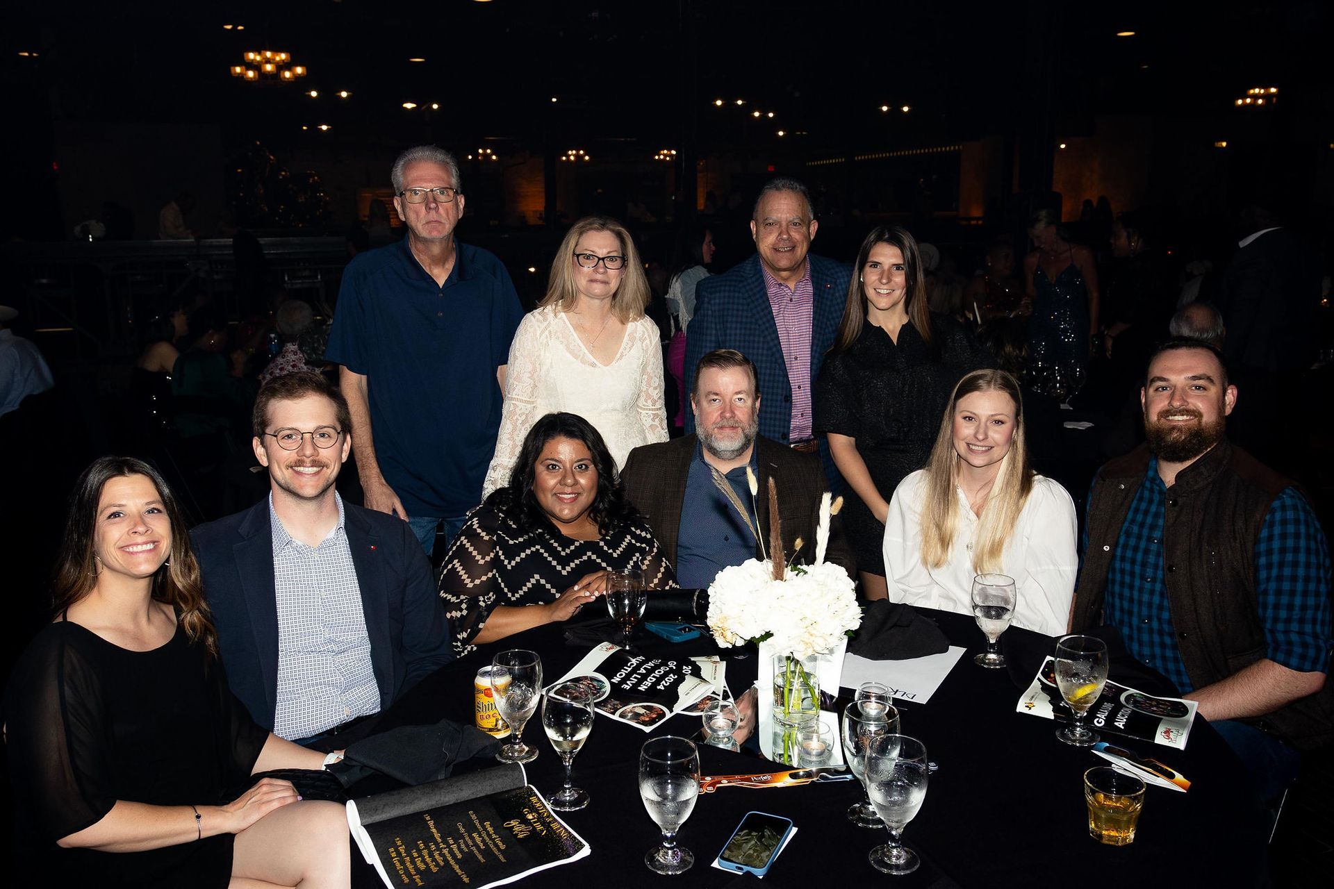 Group of people seated at a dinner table, smiling. Dimly lit room, black tablecloth, floral centerpiece.