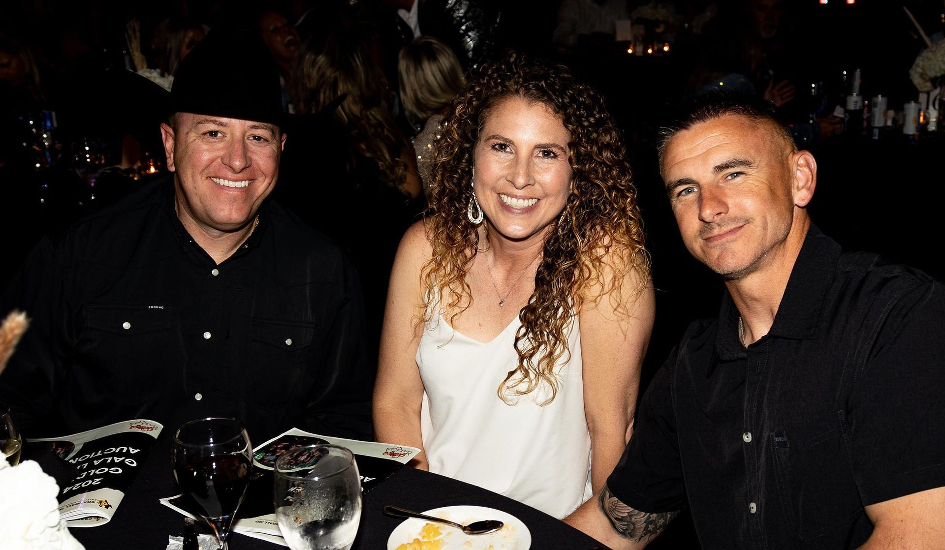 Three people smiling at a table: woman in white, men in black, dark setting.