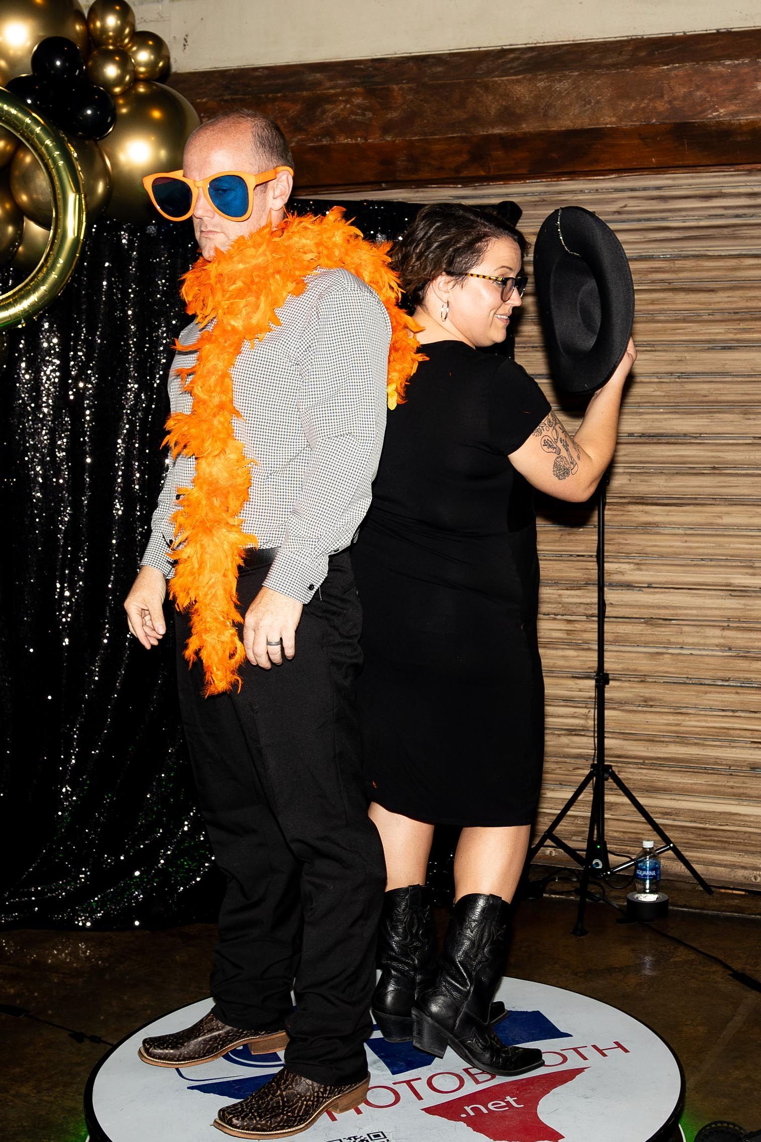 Two people pose at a photo booth; man with orange boa and shades, woman with hat, against black curtain.