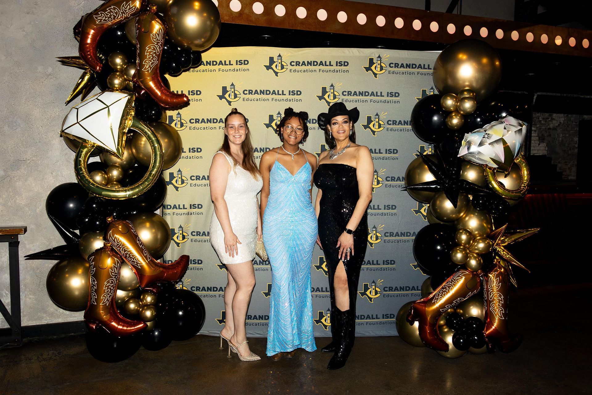 Three women pose in front of a branded backdrop with balloon decorations. One woman wears a white dress, the other a blue sequined gown, and the last in a black dress and cowboy hat.