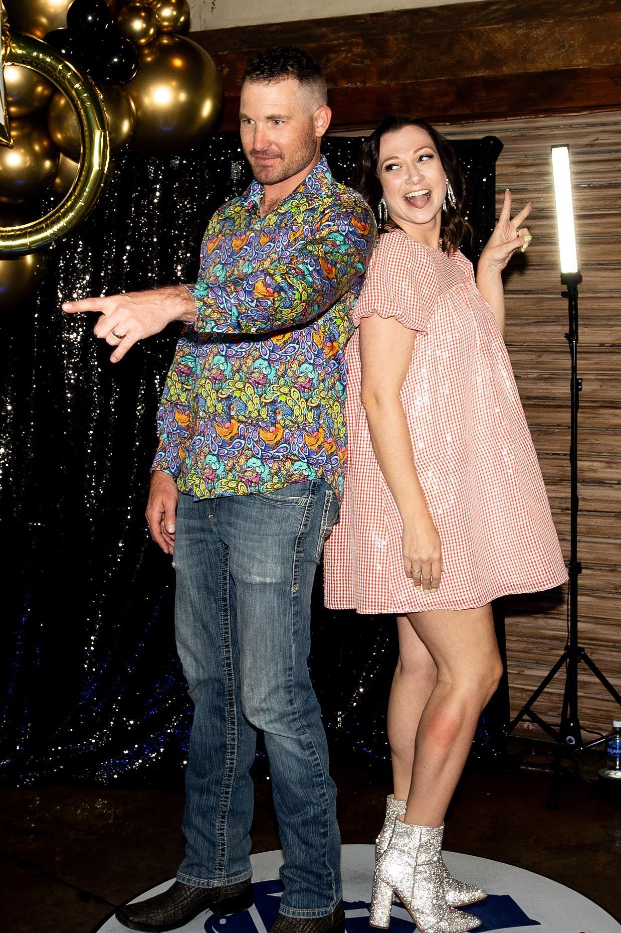 Man and woman posing, back to back. Man points left, woman makes peace sign. Both smile. Backdrop with gold balloons and lights.