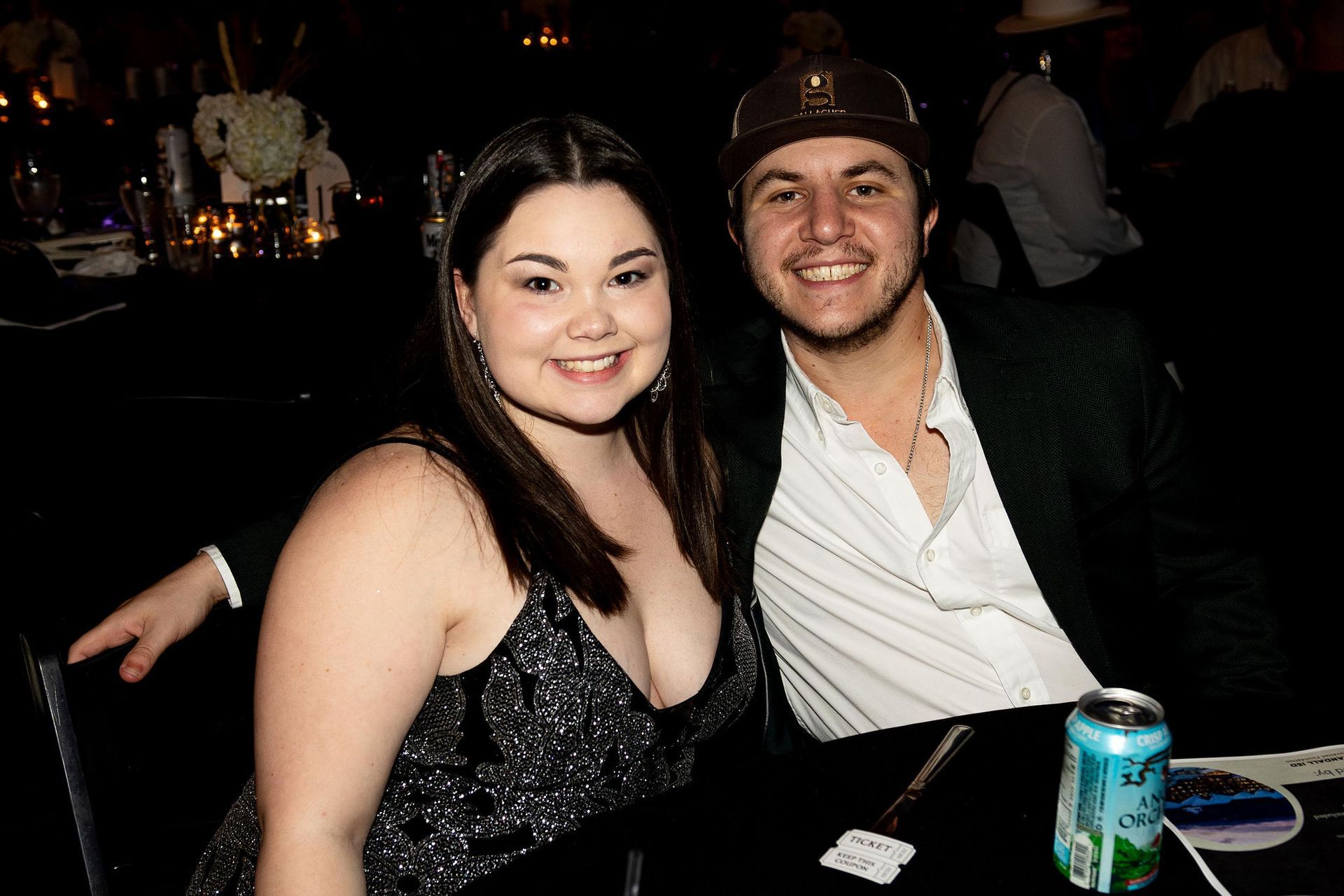 Woman in sparkly dress and man in cap smile at camera. Sitting at a table.