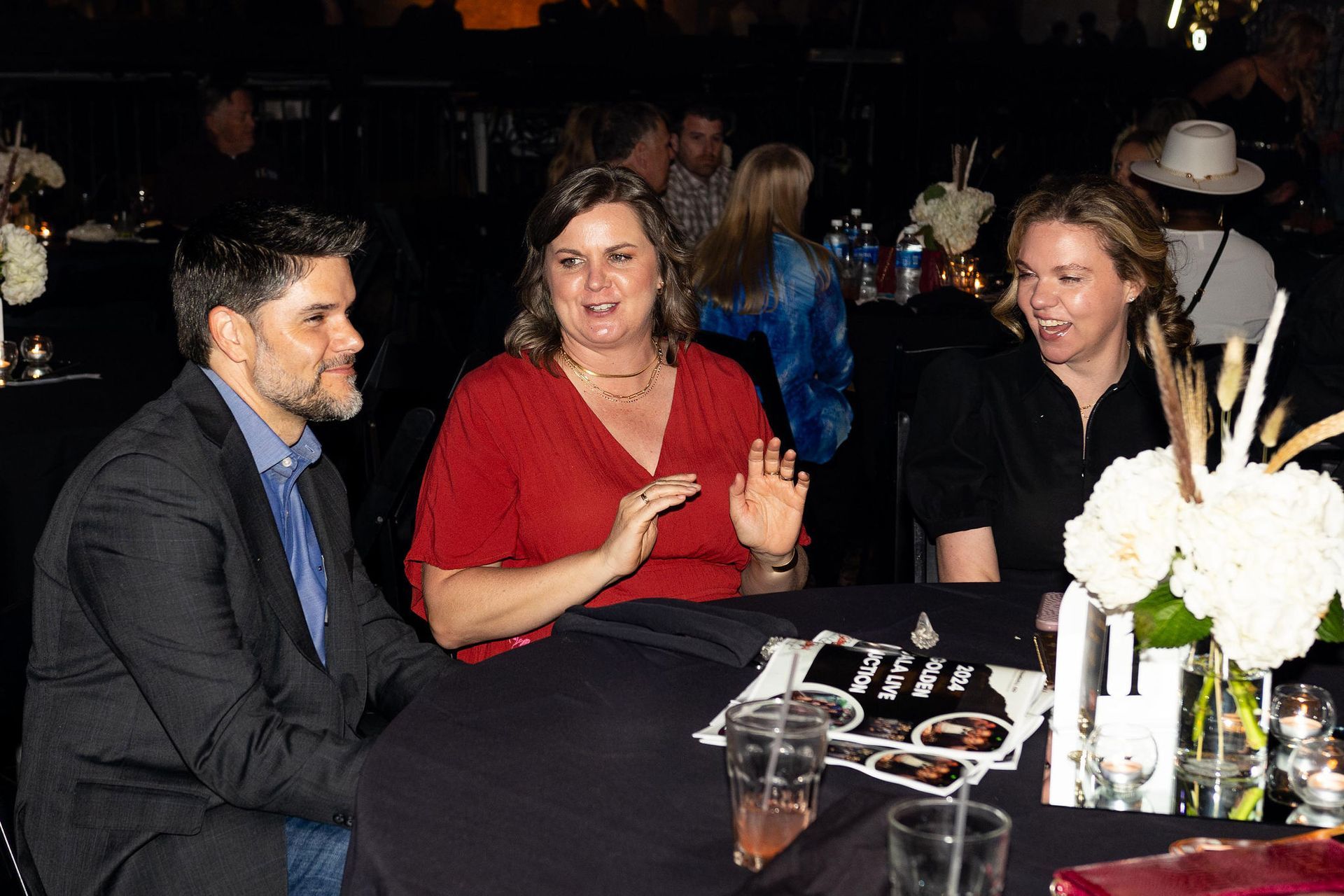 Three people at a table: a man, a woman in red gesturing, and a woman in black smiling. Flowers and drinks.