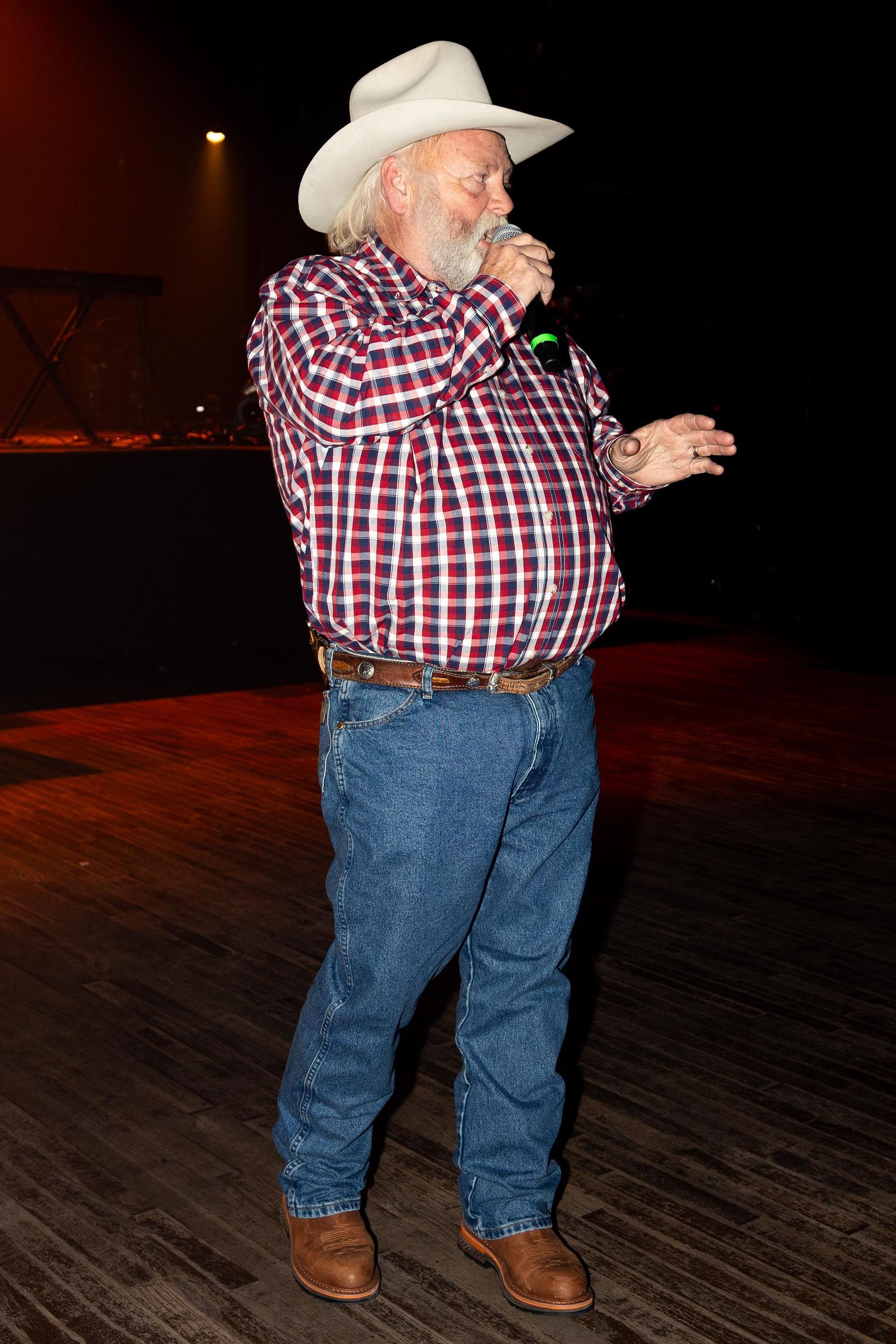 Man in cowboy hat and plaid shirt singing on stage, gesturing with one hand.