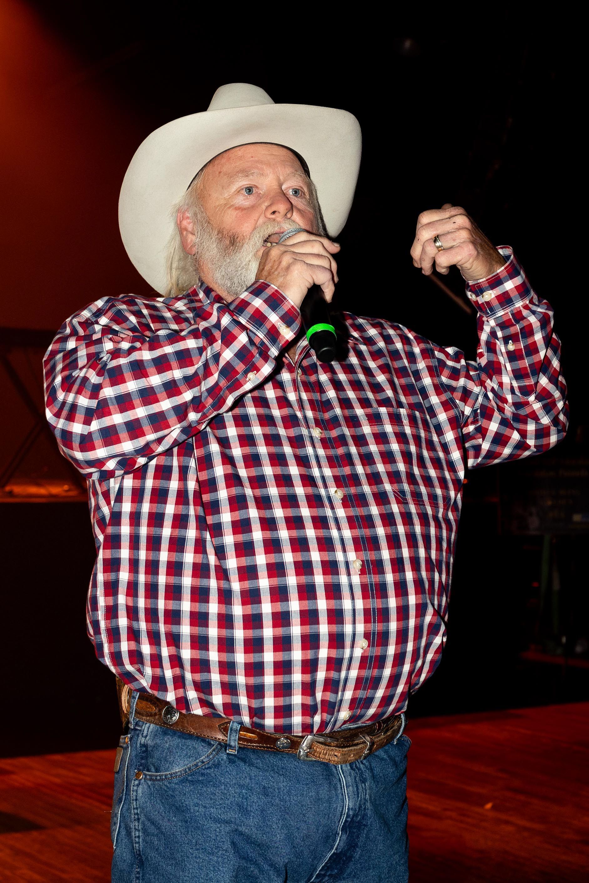 Man in cowboy hat and plaid shirt sings into a microphone on stage.