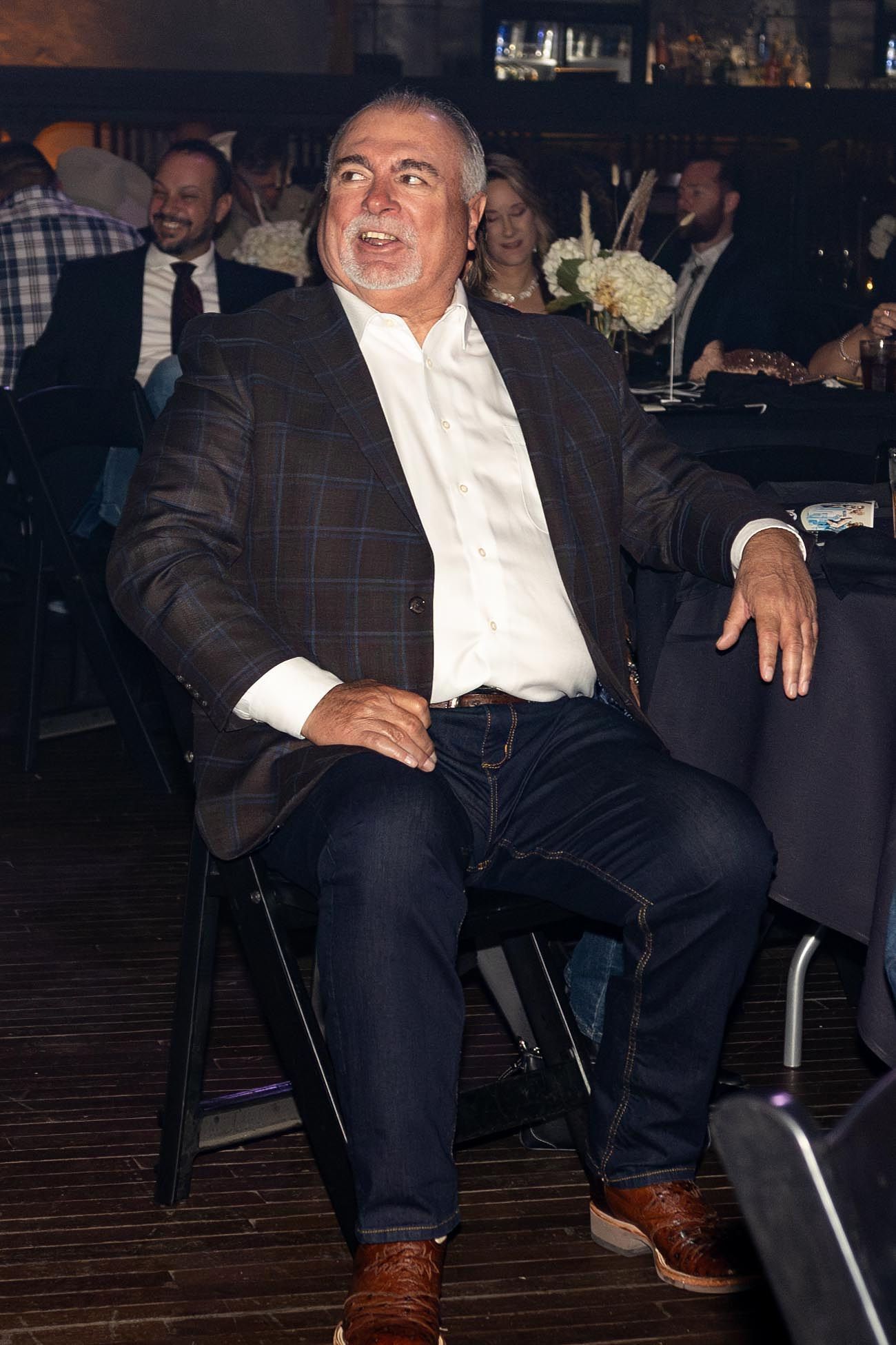 Man in a brown plaid blazer, white shirt, and jeans sits at a table, smiling.