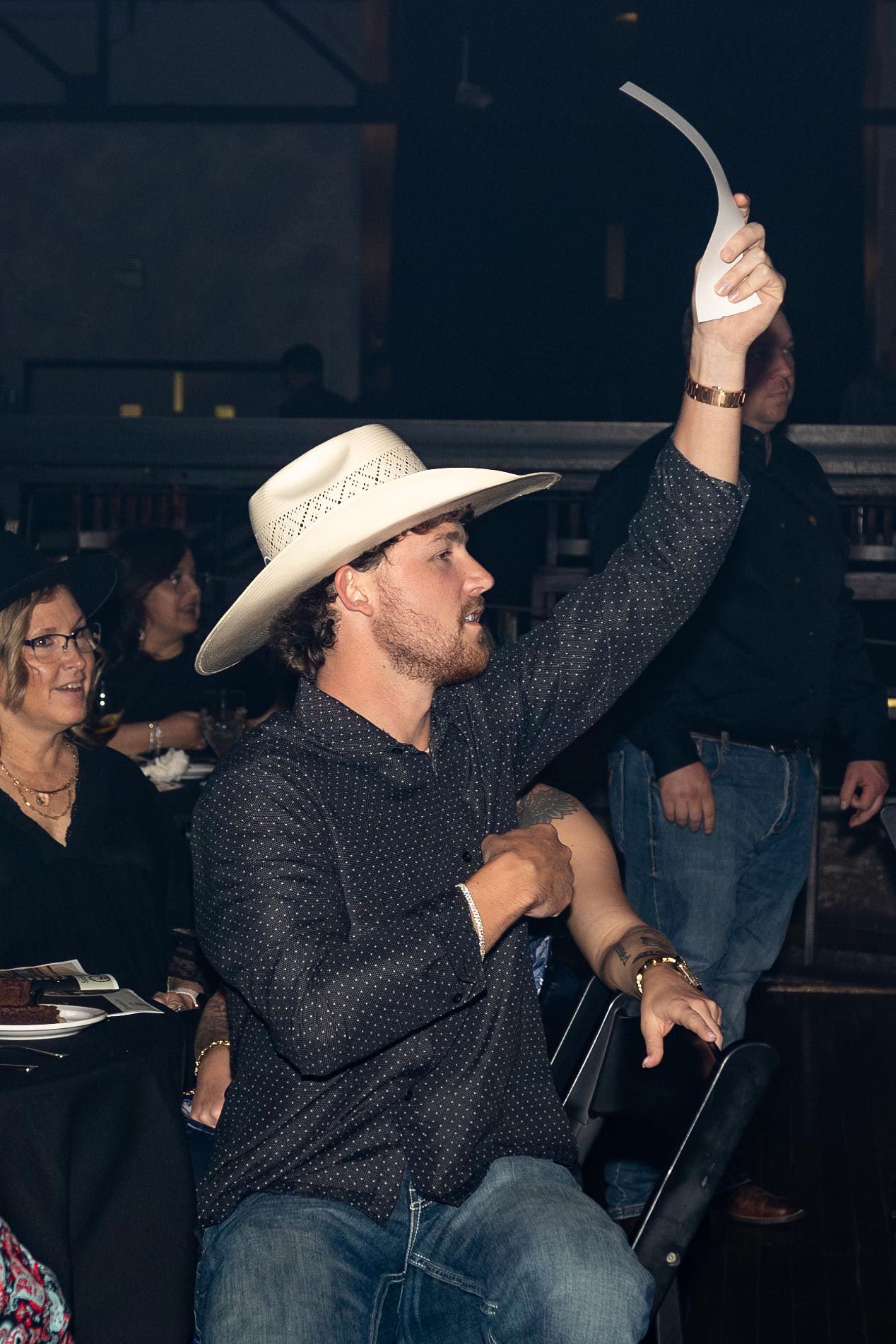 Man in cowboy hat raises a paddle at an auction; dark shirt, seated, indoor setting.