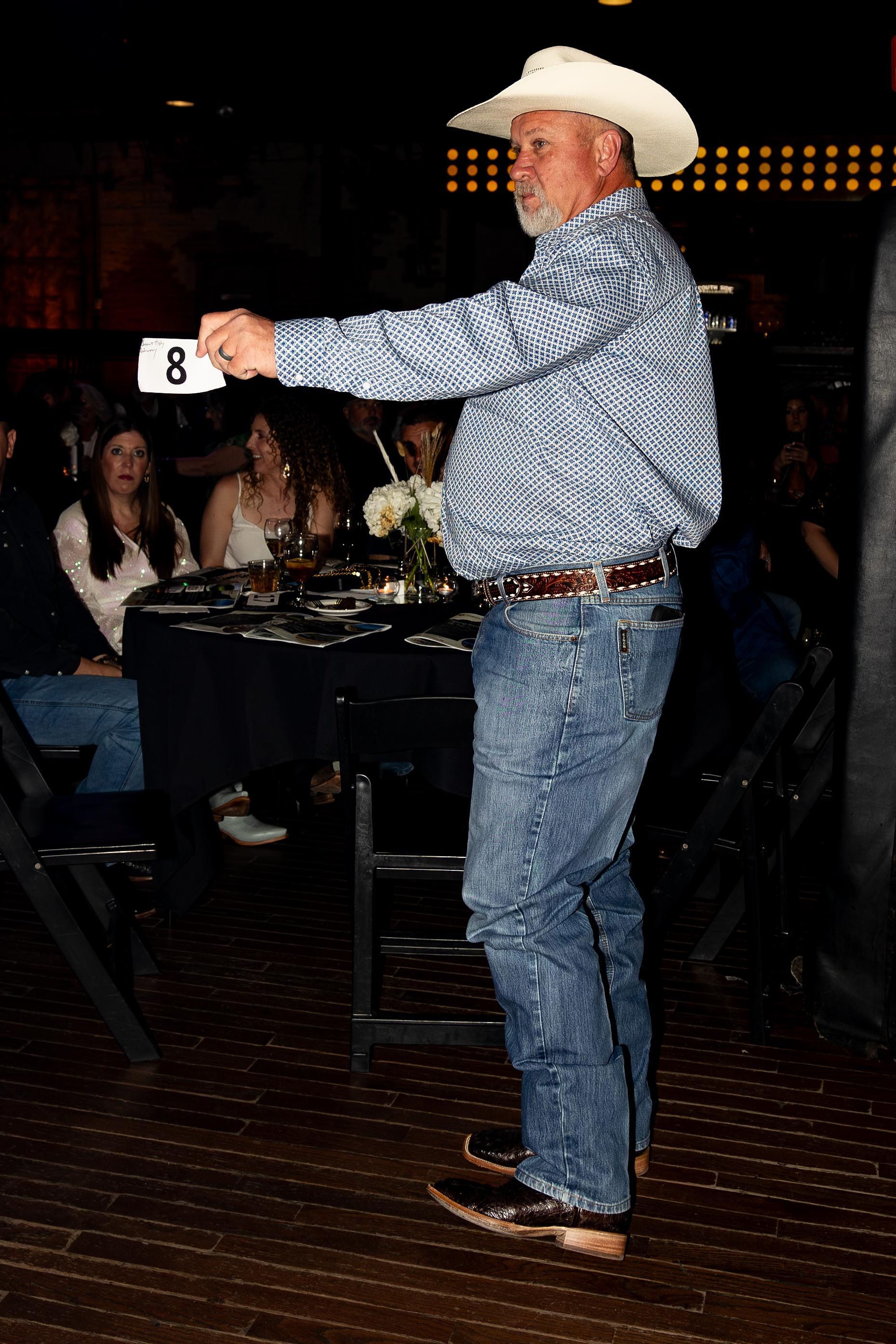 Man in cowboy hat holds number 8 at a seated event. He wears jeans, patterned shirt, and boots.