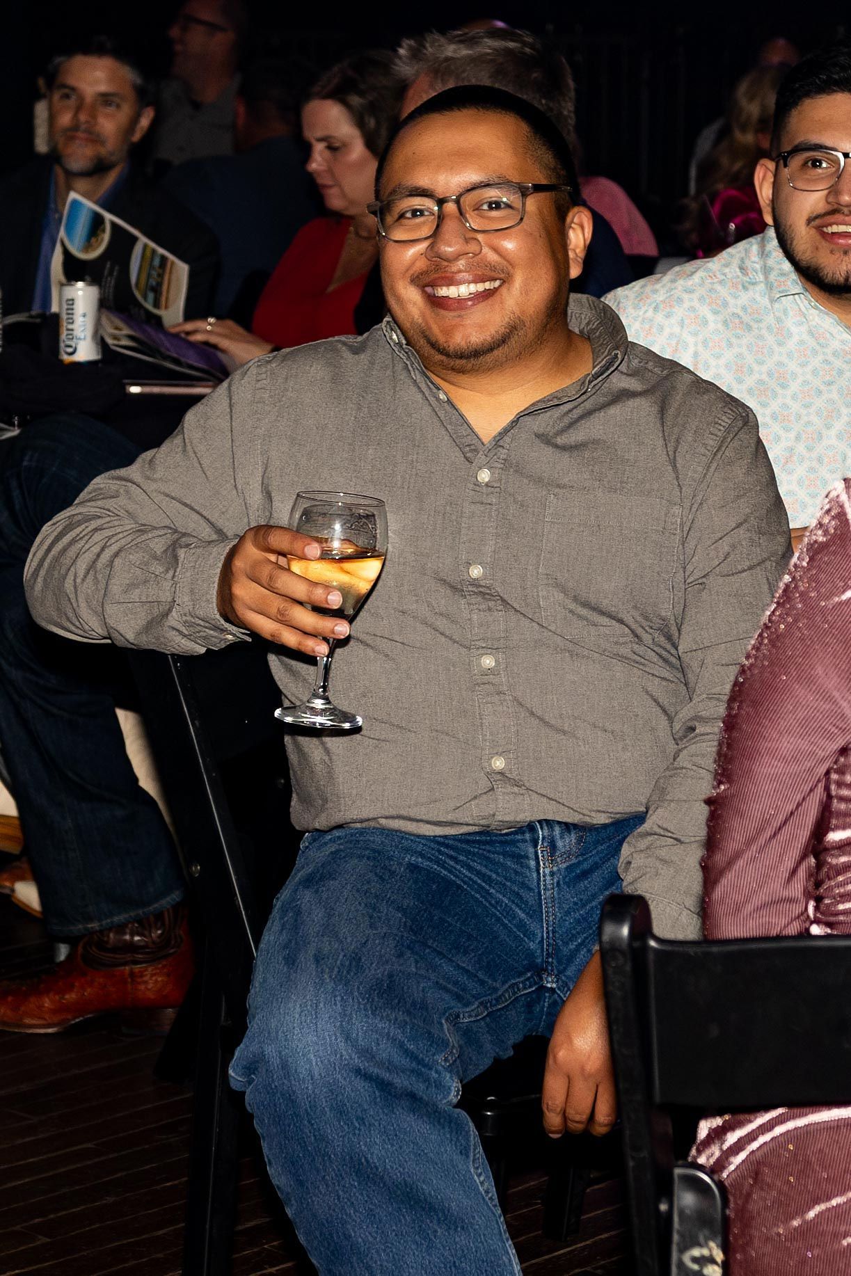Man with glasses smiles, holding a wine glass. He is wearing a button-down shirt and jeans, sitting indoors.