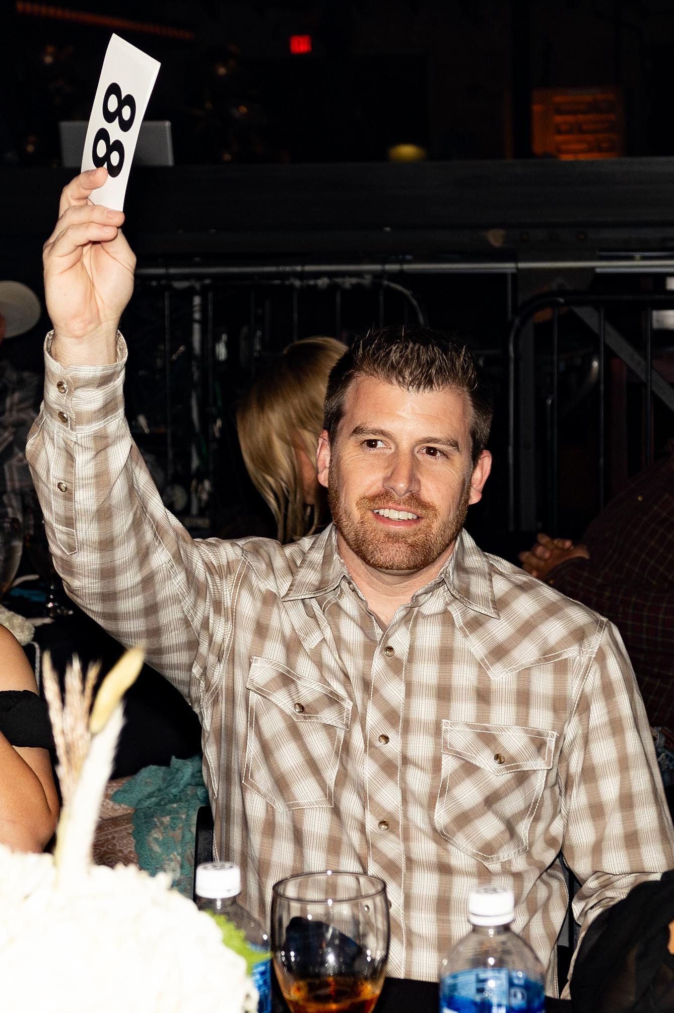 Man at auction raises bid number 88. He wears a plaid shirt and smiles.