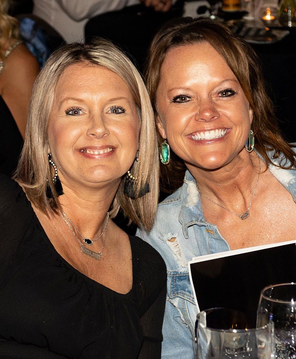 Two smiling women, one blond in black top, the other brunette in denim jacket, at a dinner.