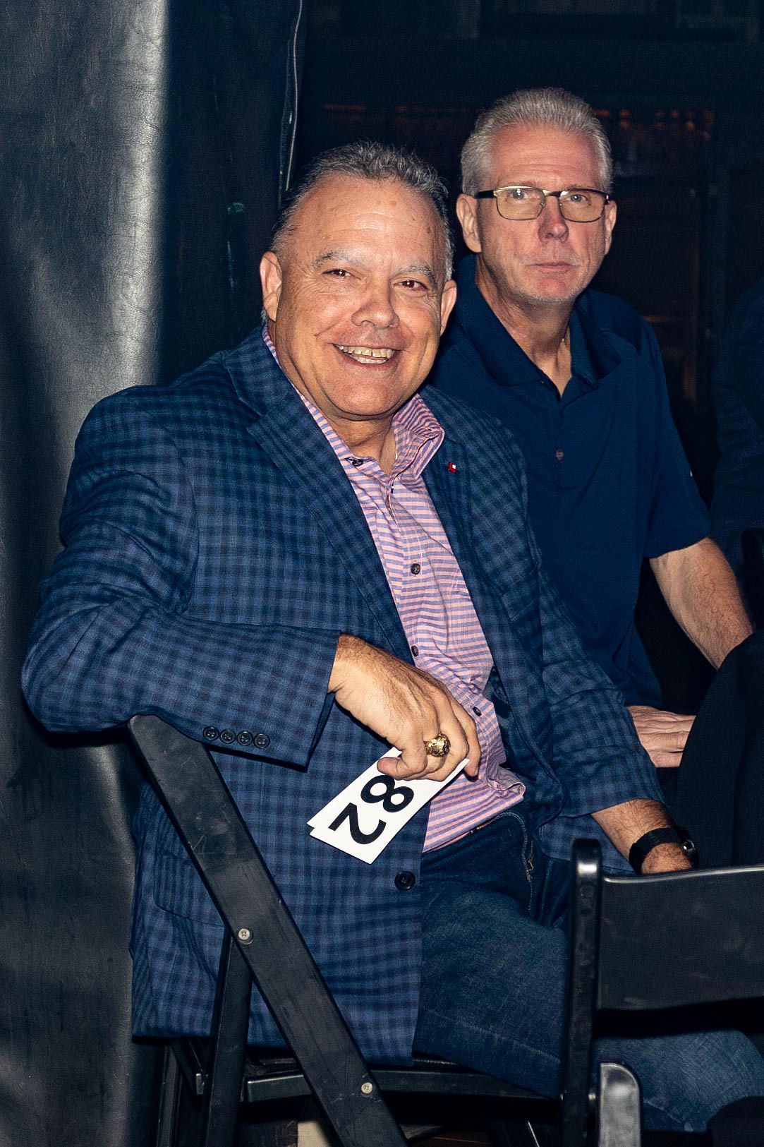 Smiling man in a blue checkered blazer with a number 82 pinned on the back of the chair. Another man behind him.