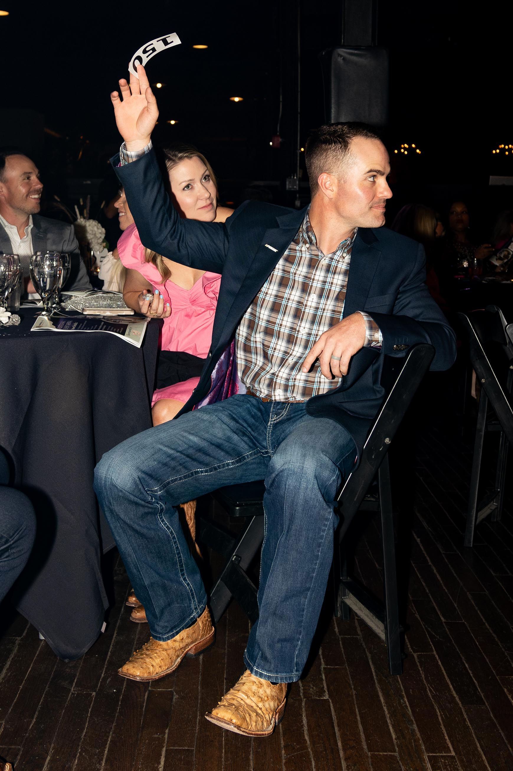 Man raising hand with cards, sitting at a table; woman watching. Indoors, dark setting.
