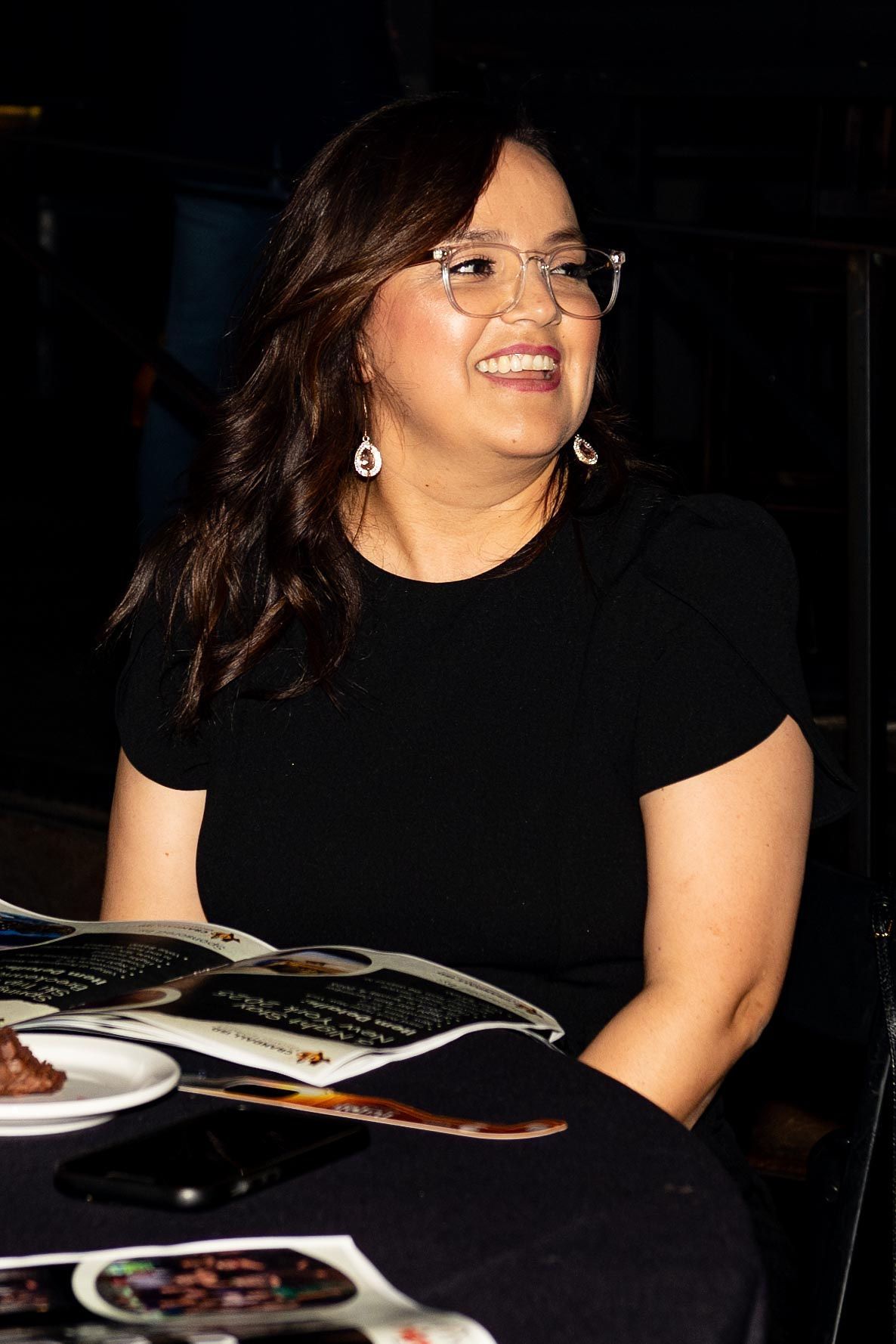 Woman with glasses smiles, sitting at a table with documents. She wears a black shirt.