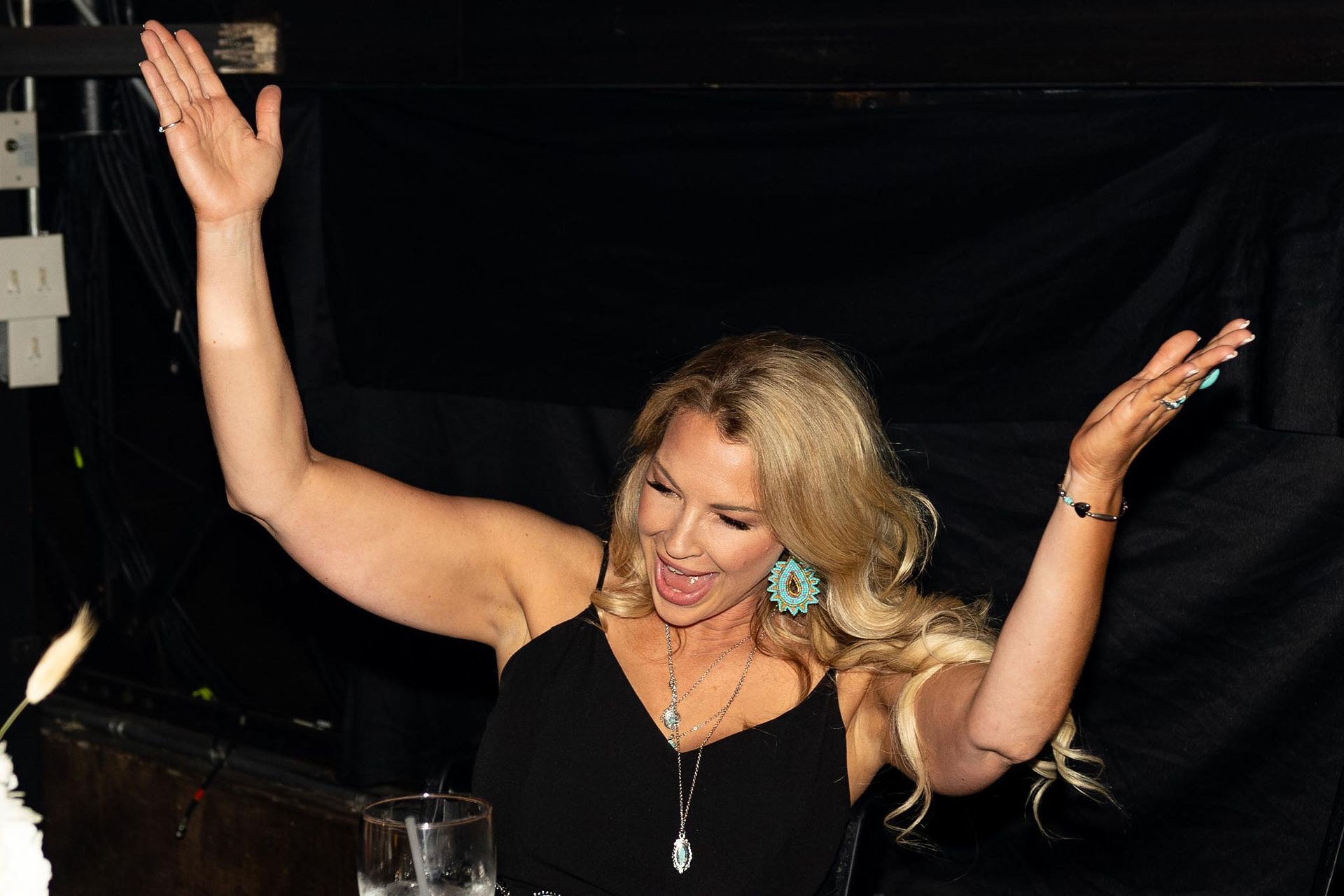 Blonde woman with arms raised celebrating. Wearing black top, turquoise earrings, and necklaces. Black background.