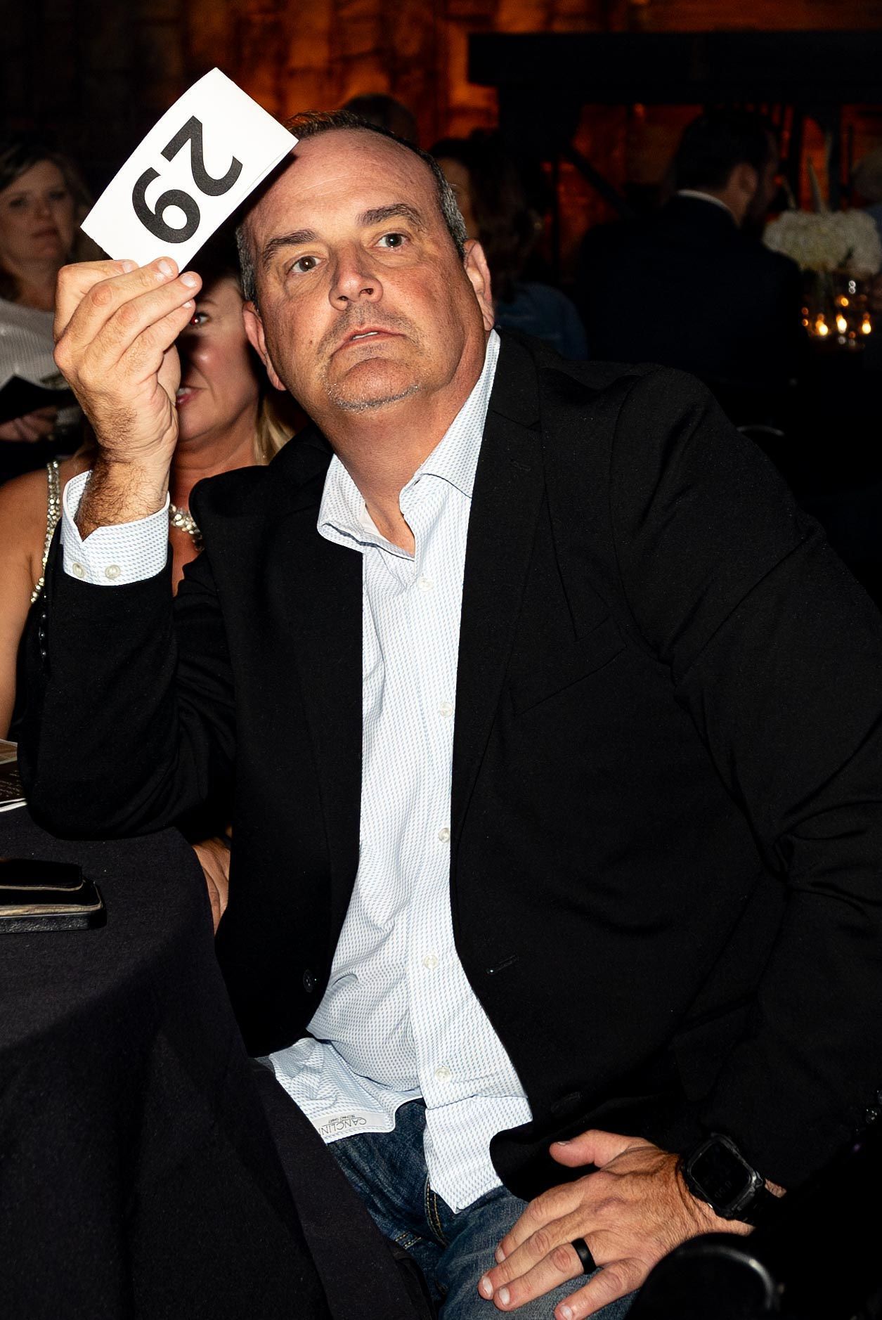 Man at auction, holding up bid paddle with number 29. Wearing black blazer, light blue patterned shirt, and jeans.