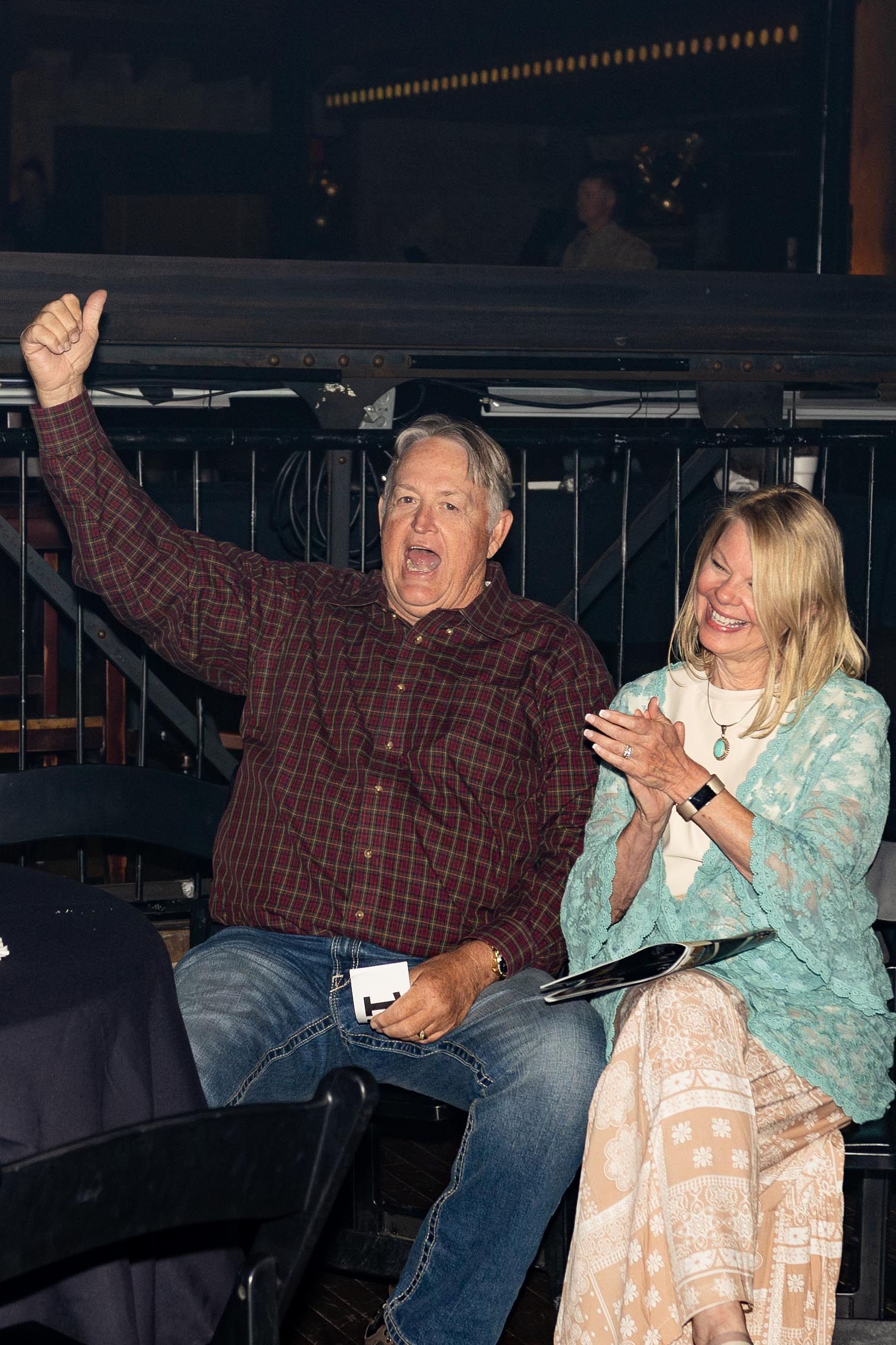 Man in plaid shirt raises thumb, woman claps beside him, both smiling. Event setting.