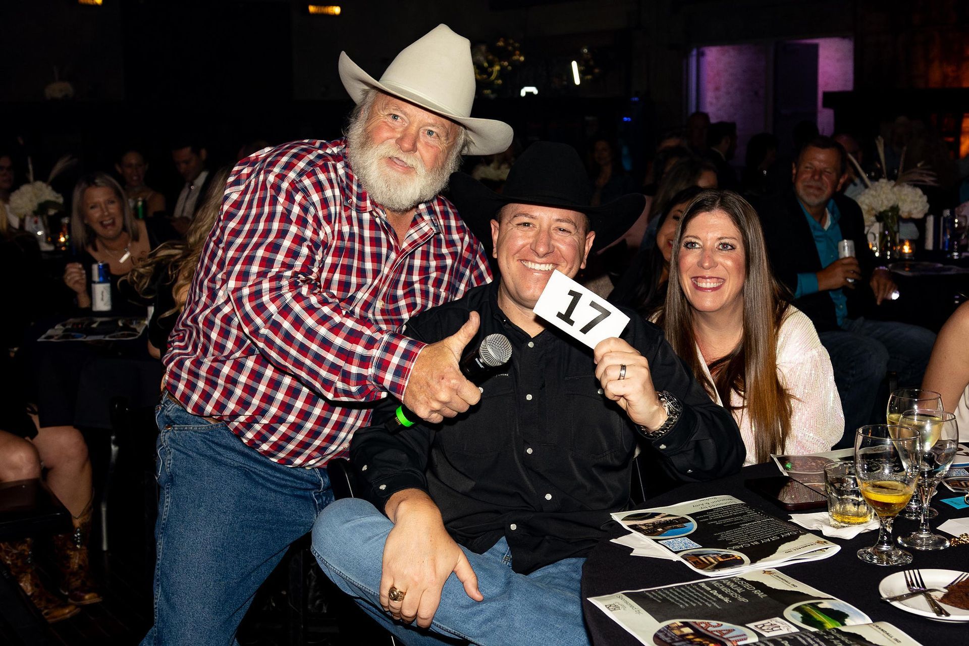 Two men and a woman at a gala. One man in a cowboy hat holds a sign; other man hugs him, all smiling.