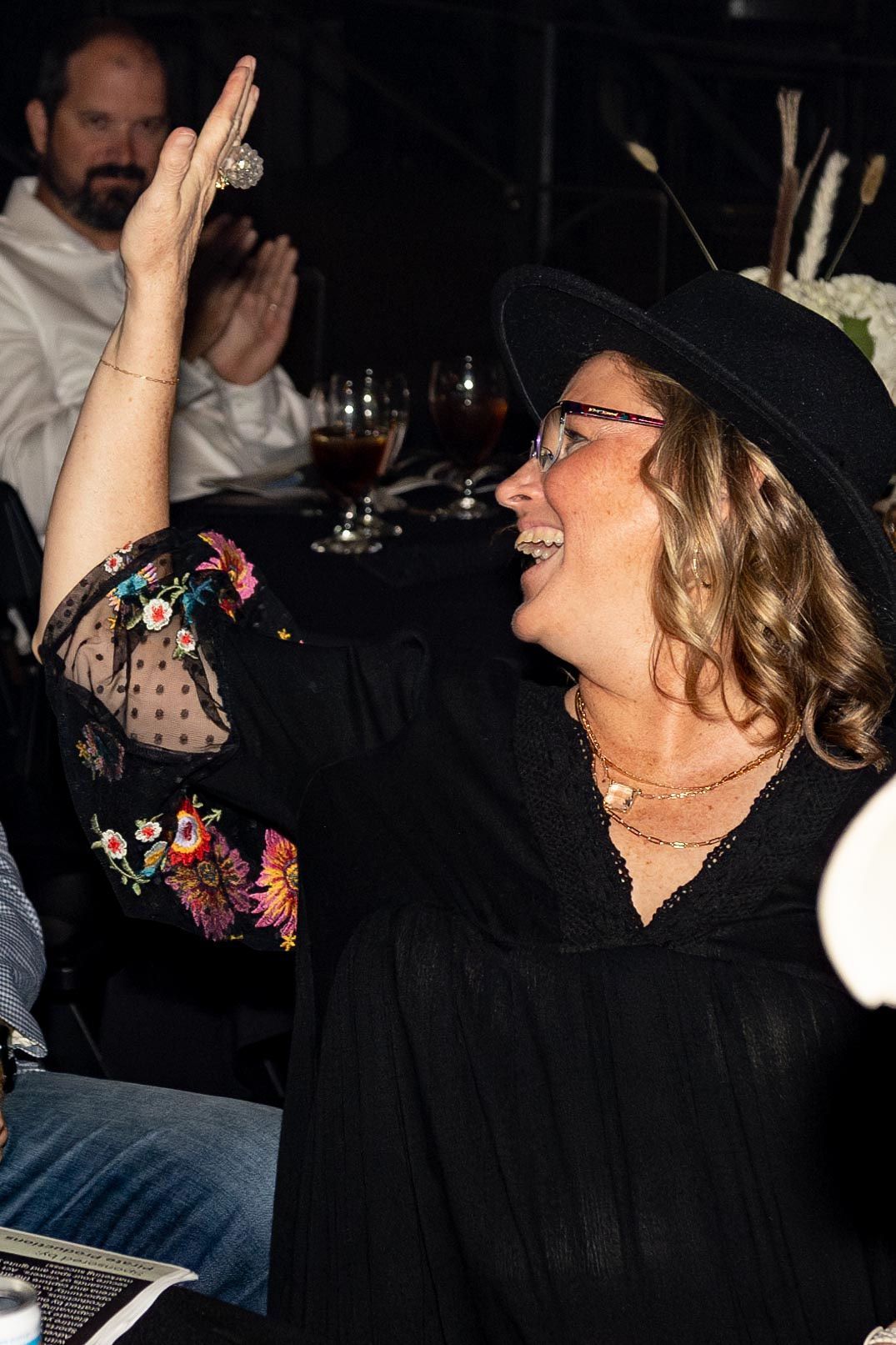 Woman in black hat and floral sleeves, smiling and raising hand, at a table.