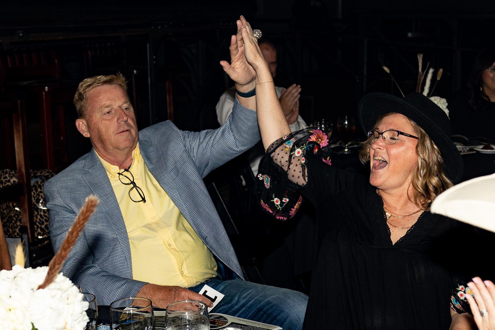 Man and woman high-fiving; man in blue blazer, woman in black hat.  Inside at an event.