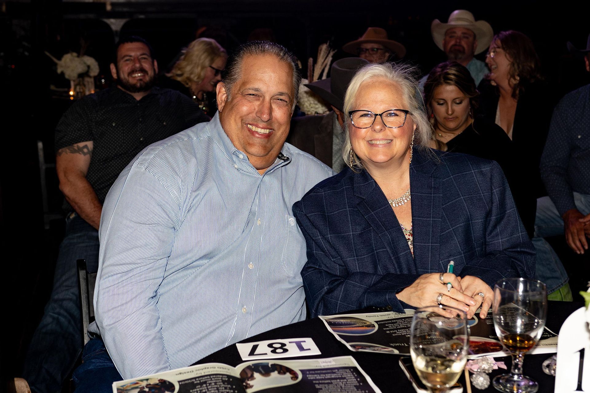Man and woman smiling at a table. They're inside, at an event. Other people in the background.