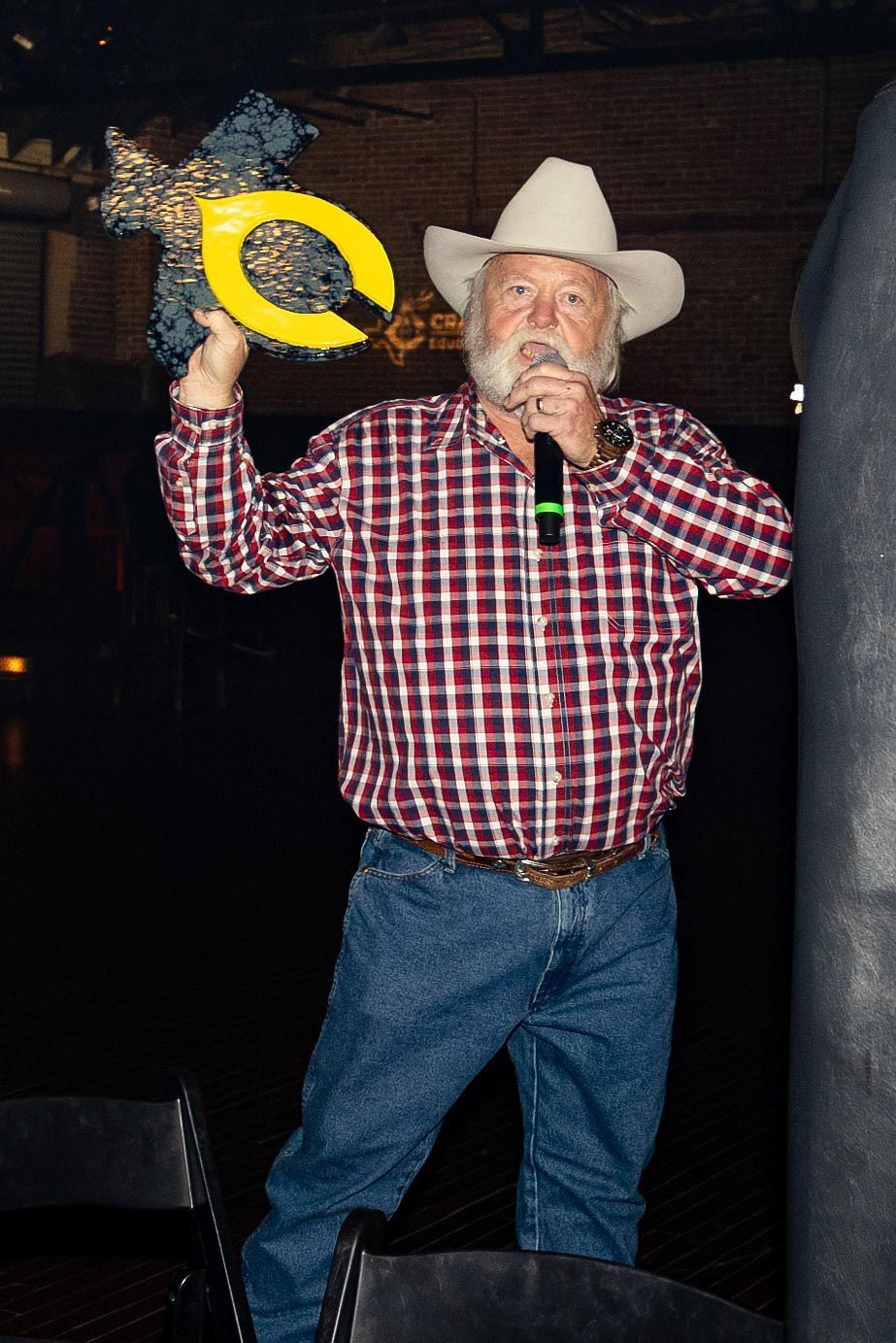 Man in cowboy hat speaking into a microphone, holding a trophy. He wears a plaid shirt and jeans.