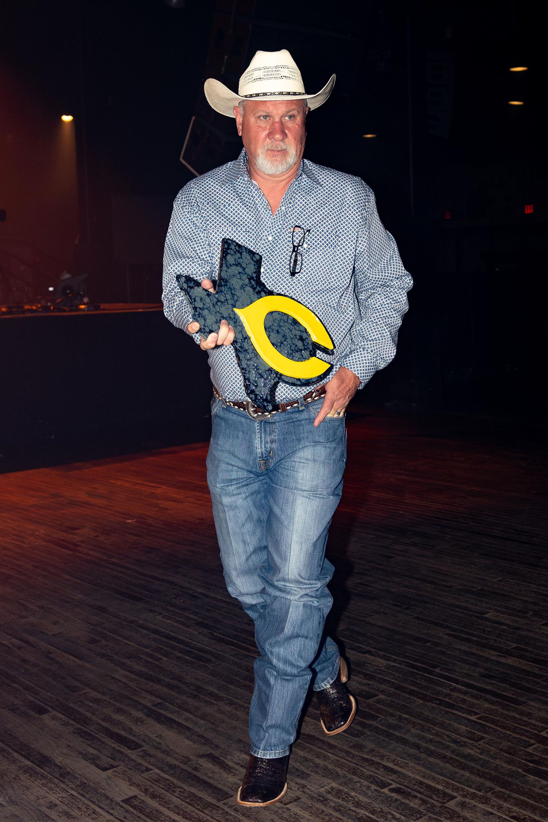 Man in cowboy hat, holding an award, walks forward. He is in a dark setting, wearing jeans and a patterned shirt.