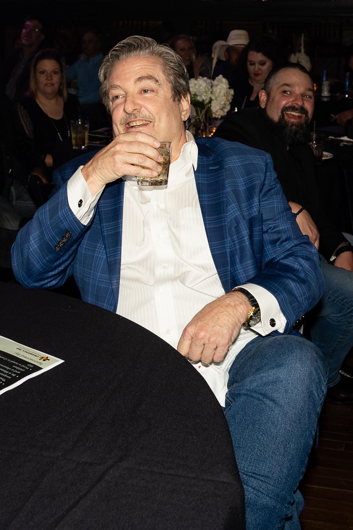Man in blue blazer drinks from a glass at a table. People in background, indoor setting.