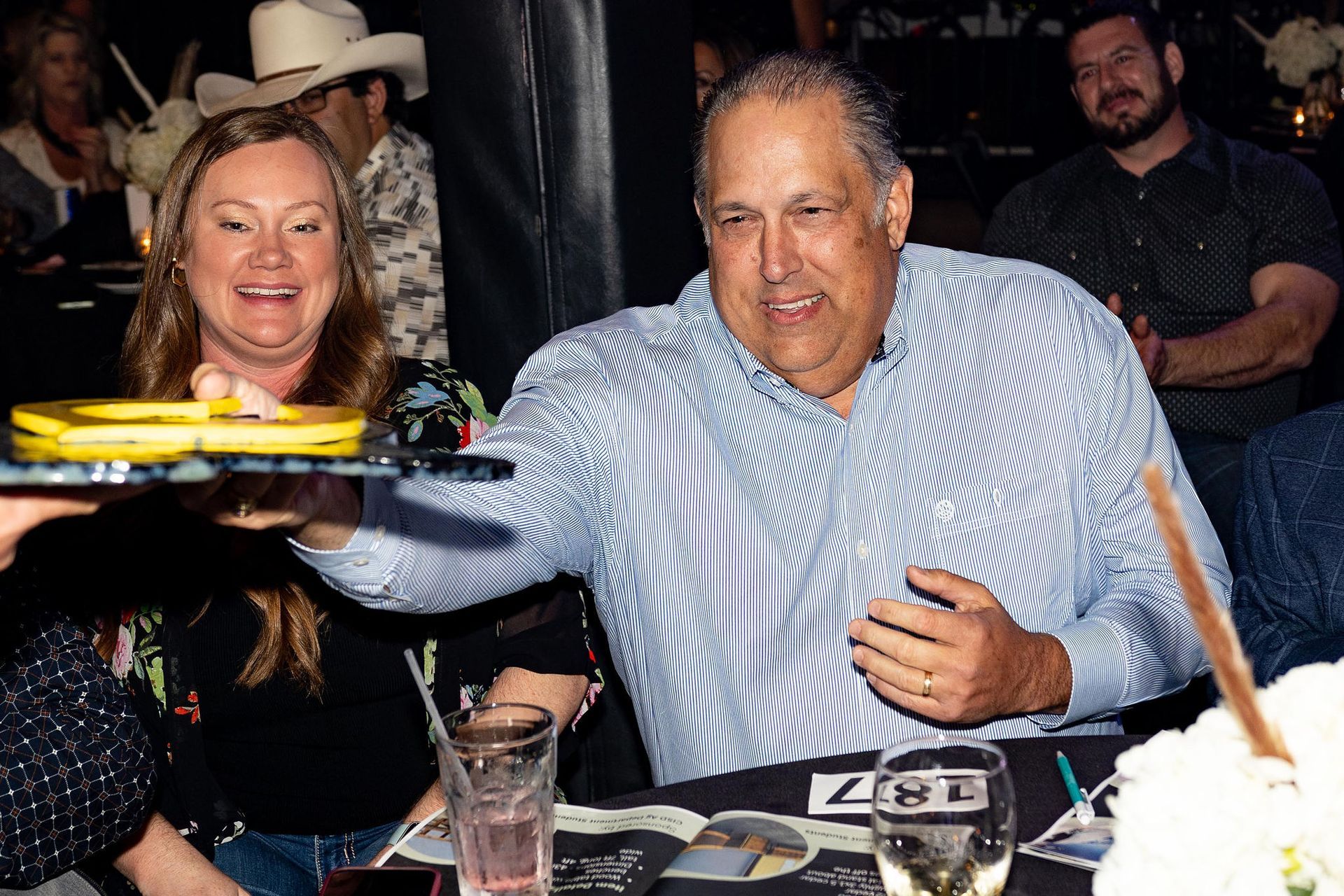 Woman and man at a table reaching for a tray with food. They smile, in a well-lit setting.