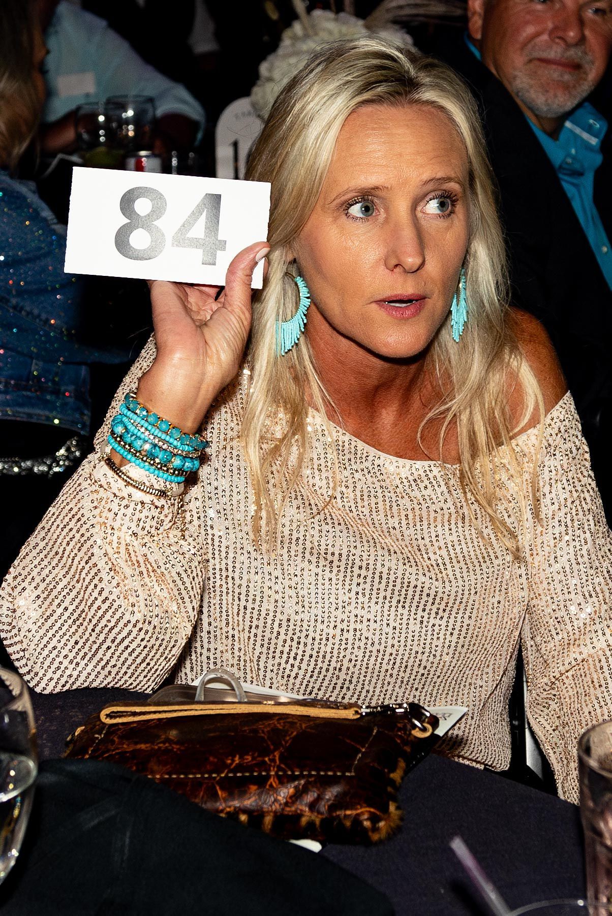 Woman at auction holding up number 84. Blonde hair, turquoise jewelry, tan sweater, dark bag.