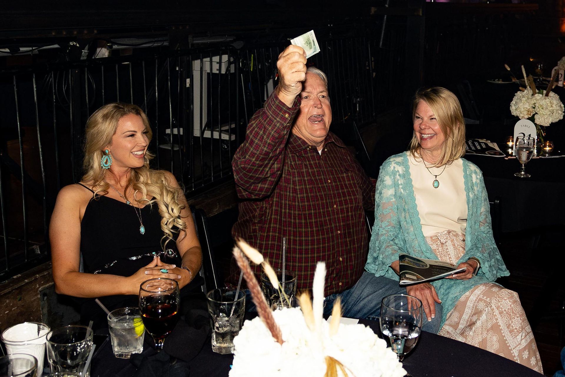 Man holding up money, flanked by two women at a table with drinks, at a party.