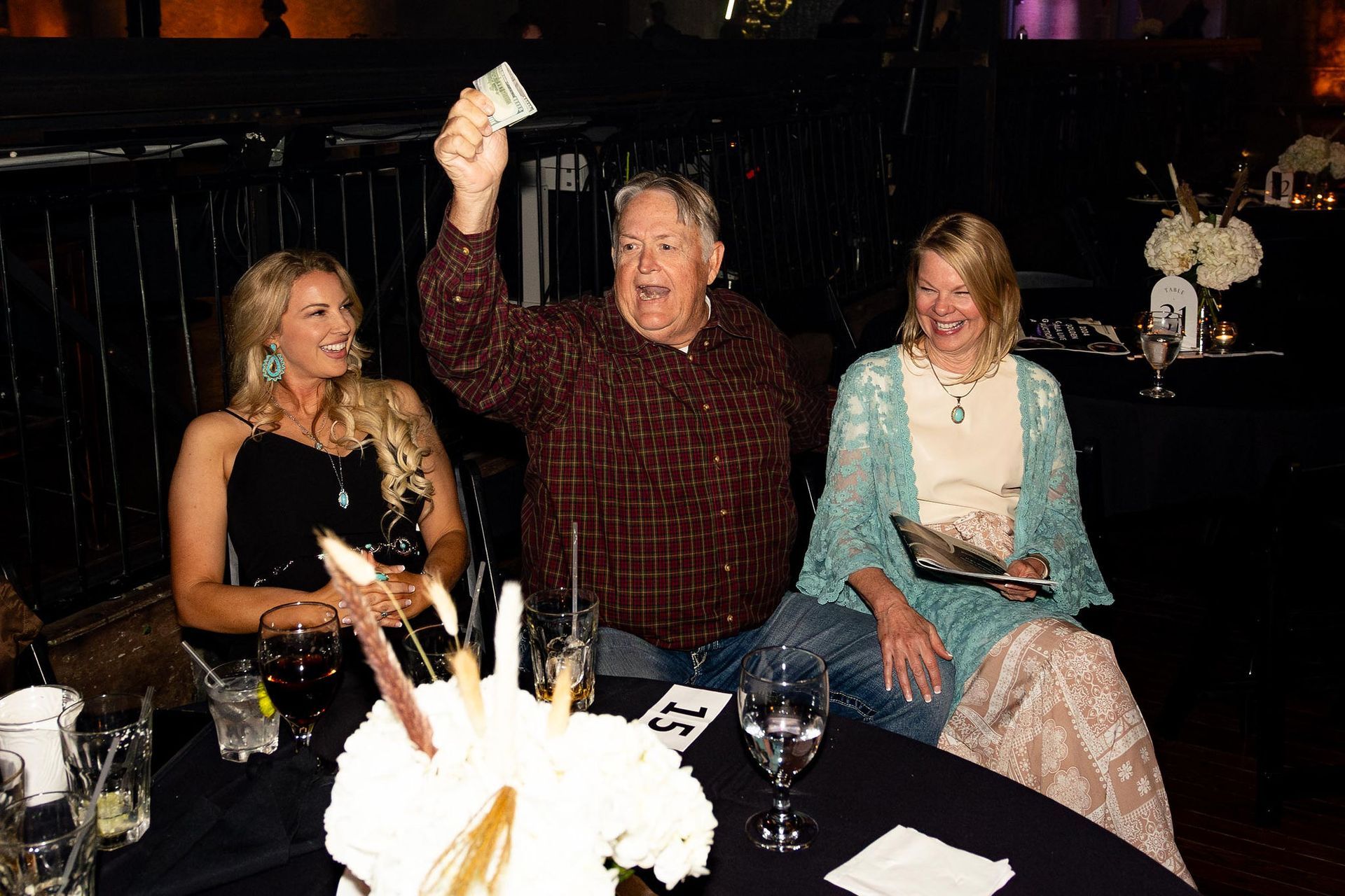 Man holding up money, excited, seated with two women at a table in a dimly lit room.
