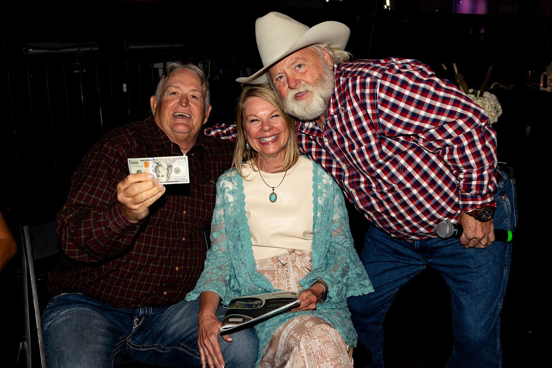 Three people smiling at a Western-themed event; one holding cash, another a microphone, and one with a turquoise necklace.