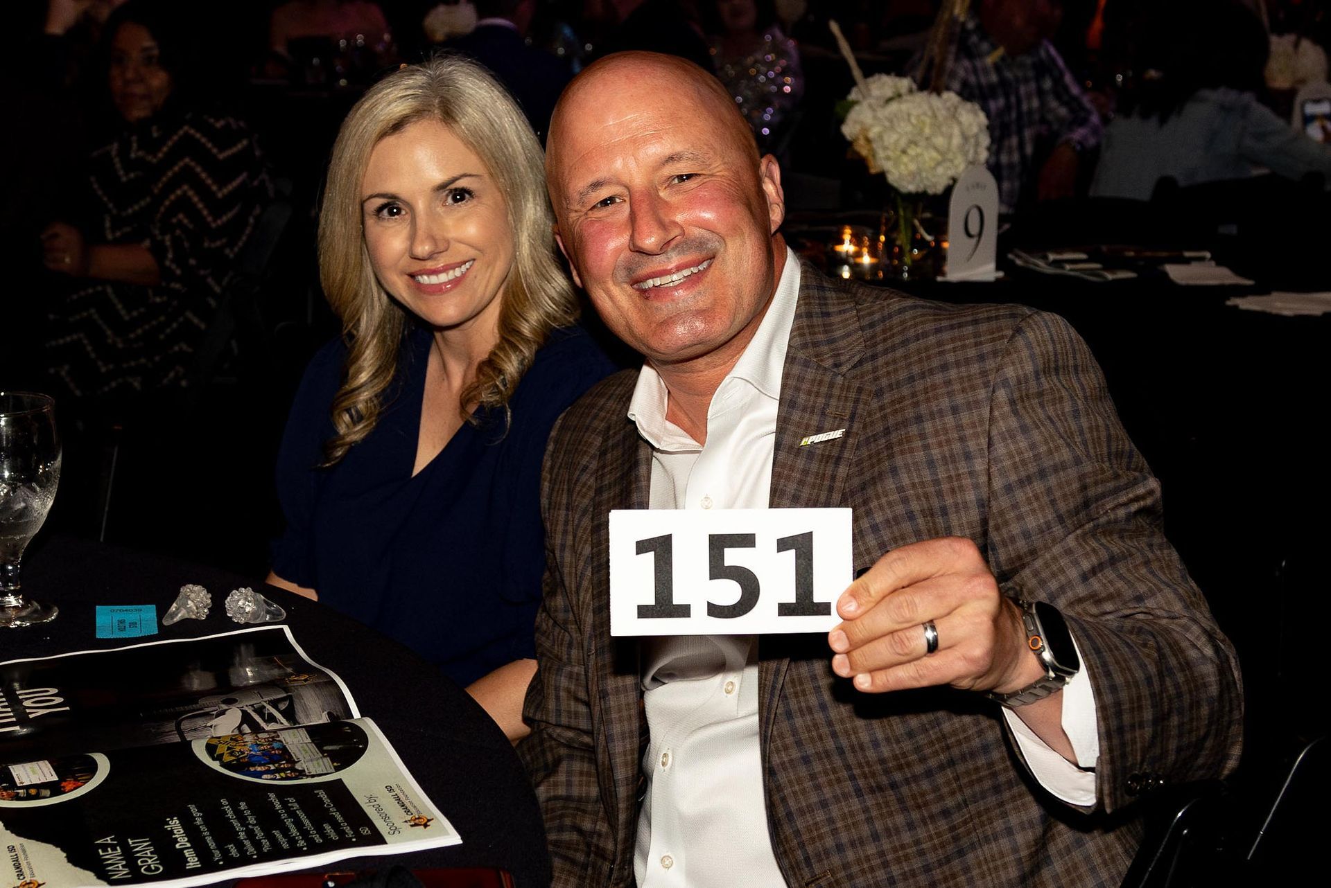 Couple at a formal event; man holds up bid number 151; woman smiles.