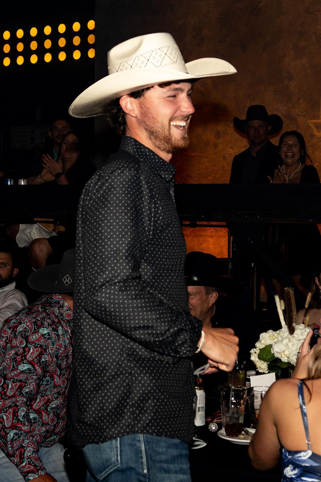 Man in cowboy hat smiles, wearing a black shirt and jeans, at an event with others in background.