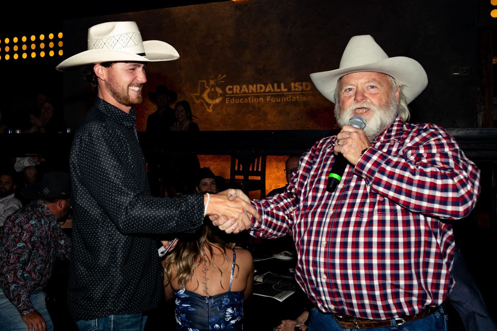 Two men in cowboy hats shaking hands at a Crandall ISD event. One holds a microphone.