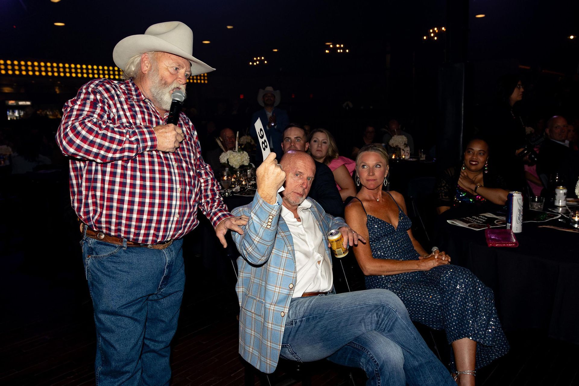 A man in a cowboy hat speaks into a mic. Two people sit nearby; a man with a guitar, and a woman.