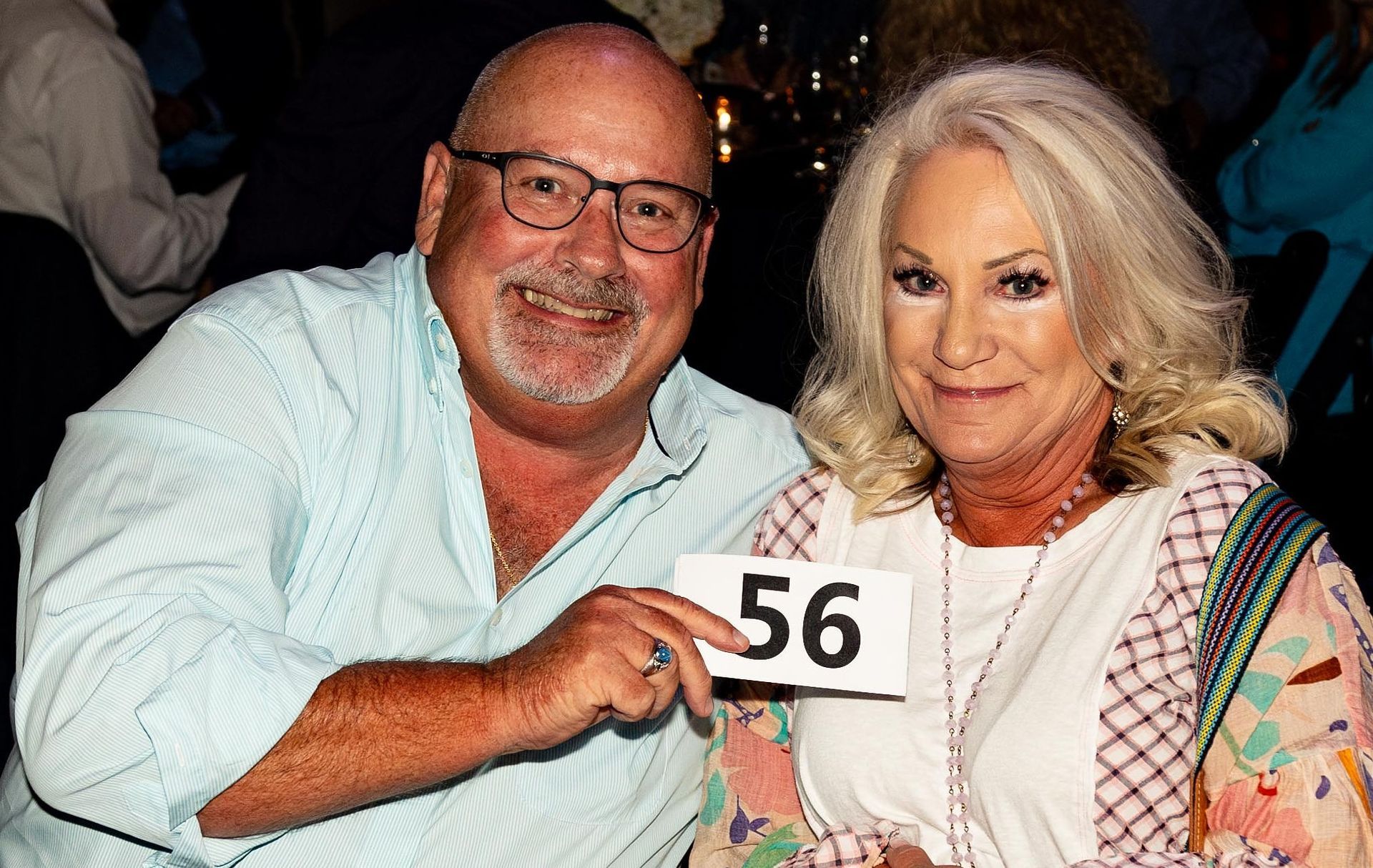 Man and woman holding a number 56 sign, smiling. Both are seated indoors.