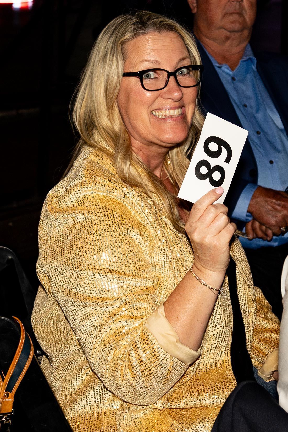 Woman with blonde hair, wearing glasses, holding auction paddle with number 89; smiling.