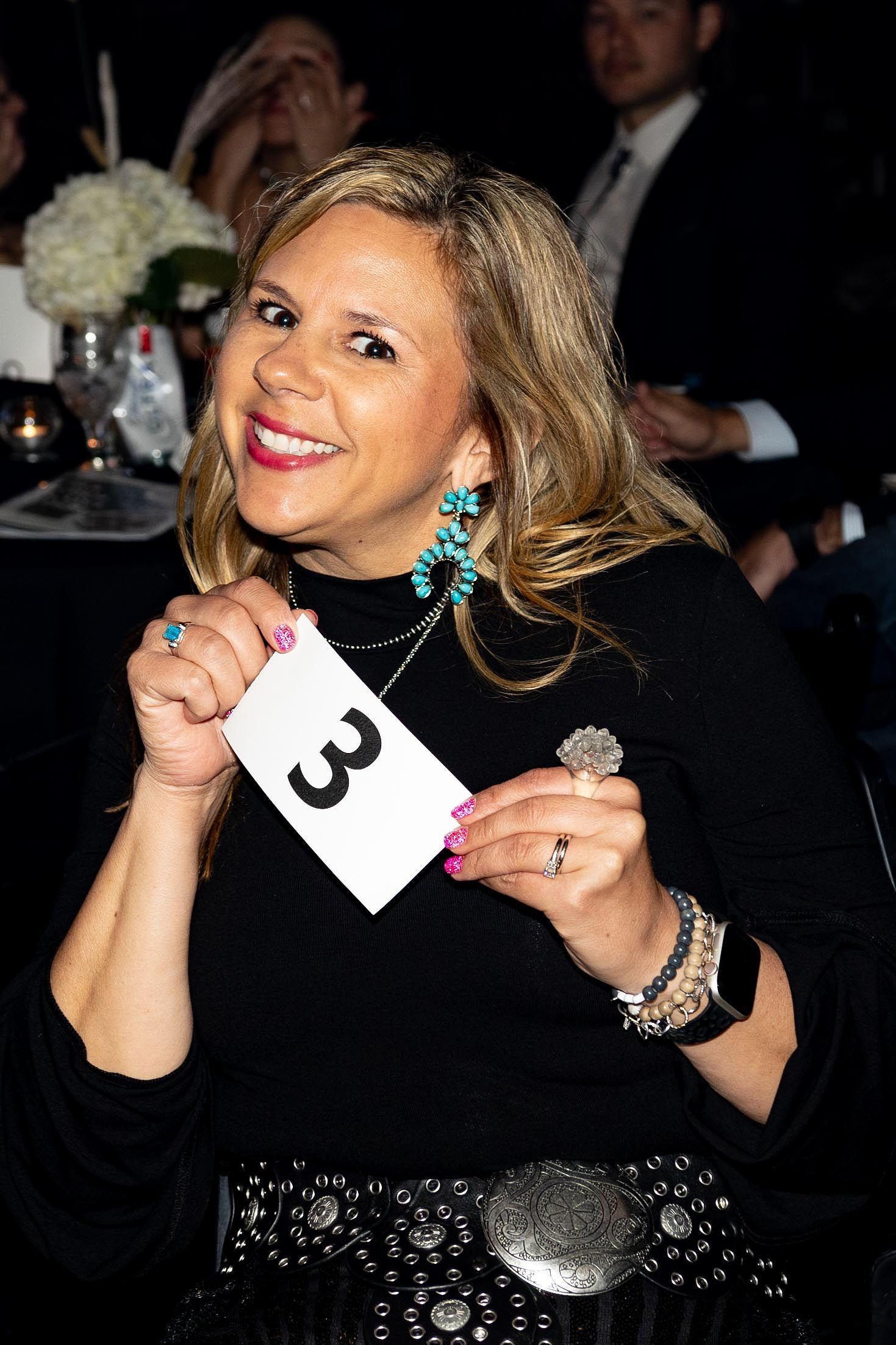 Woman smiling, holding card numbered 3. Wearing turquoise jewelry, black top, and belt.