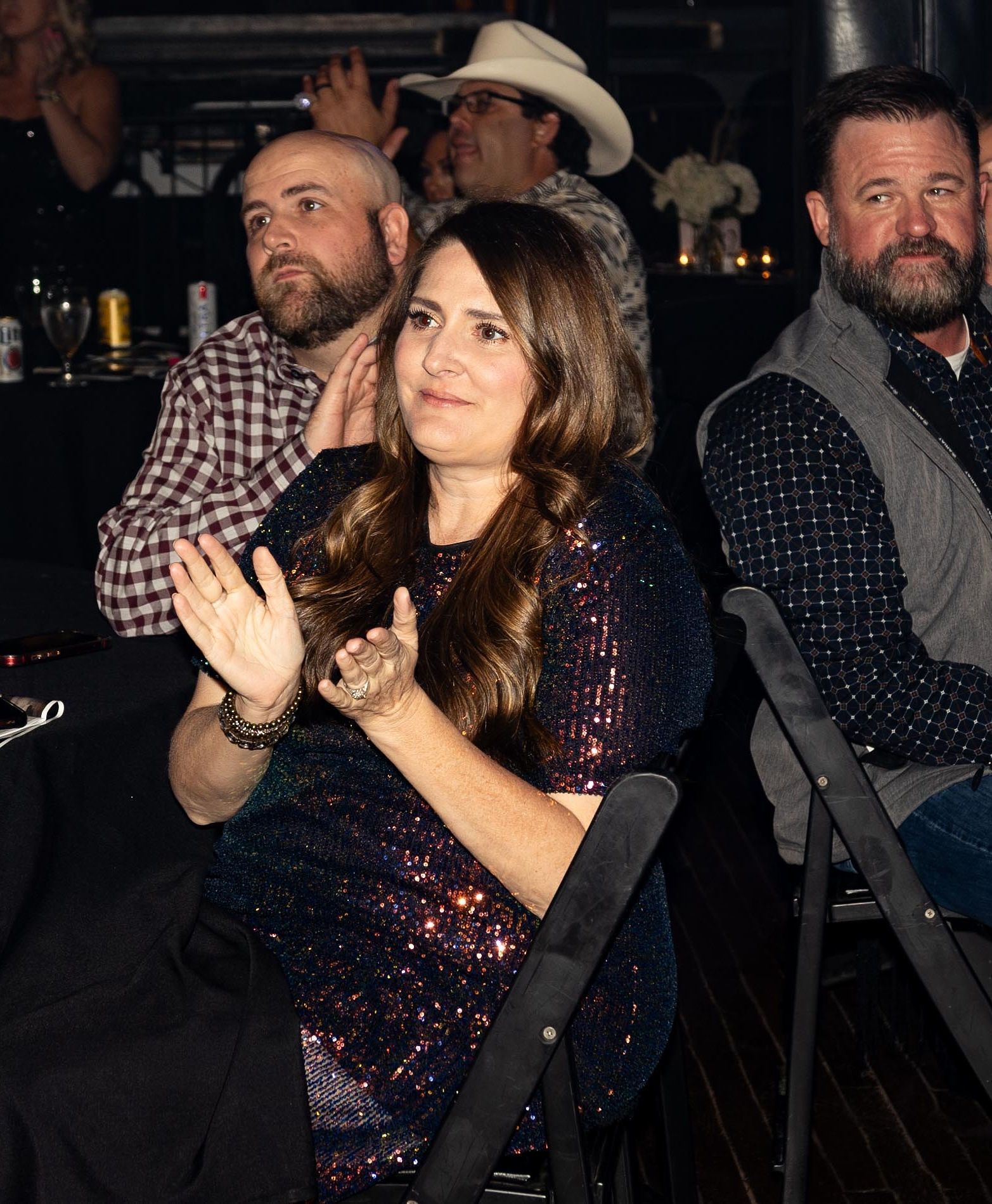 Woman in a sequin dress applauds at an event; two men look on, one wearing a cowboy hat.