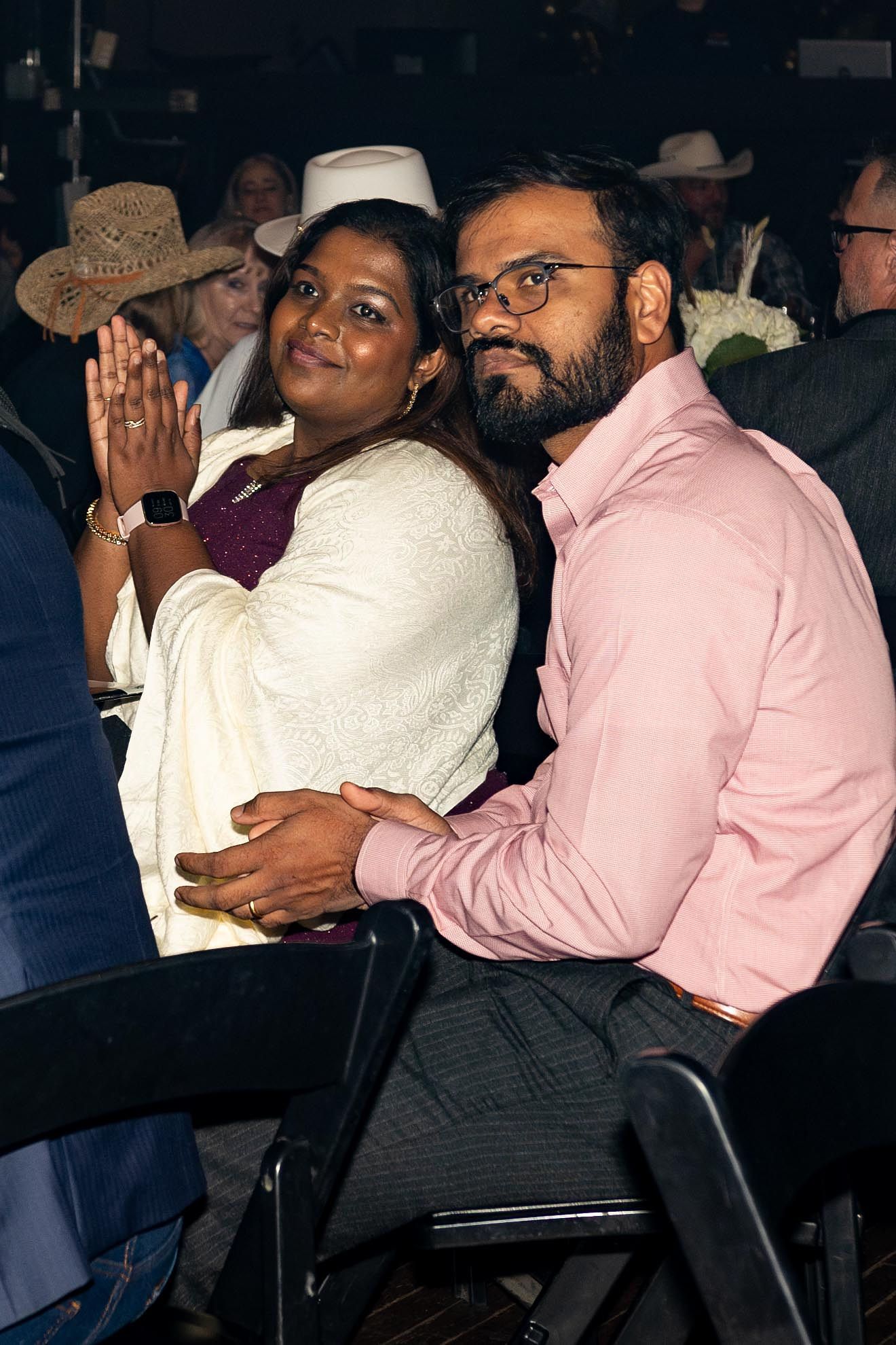 Couple seated indoors, smiling. Woman claps hands, wearing a white shawl. Man wears pink shirt, has a beard.