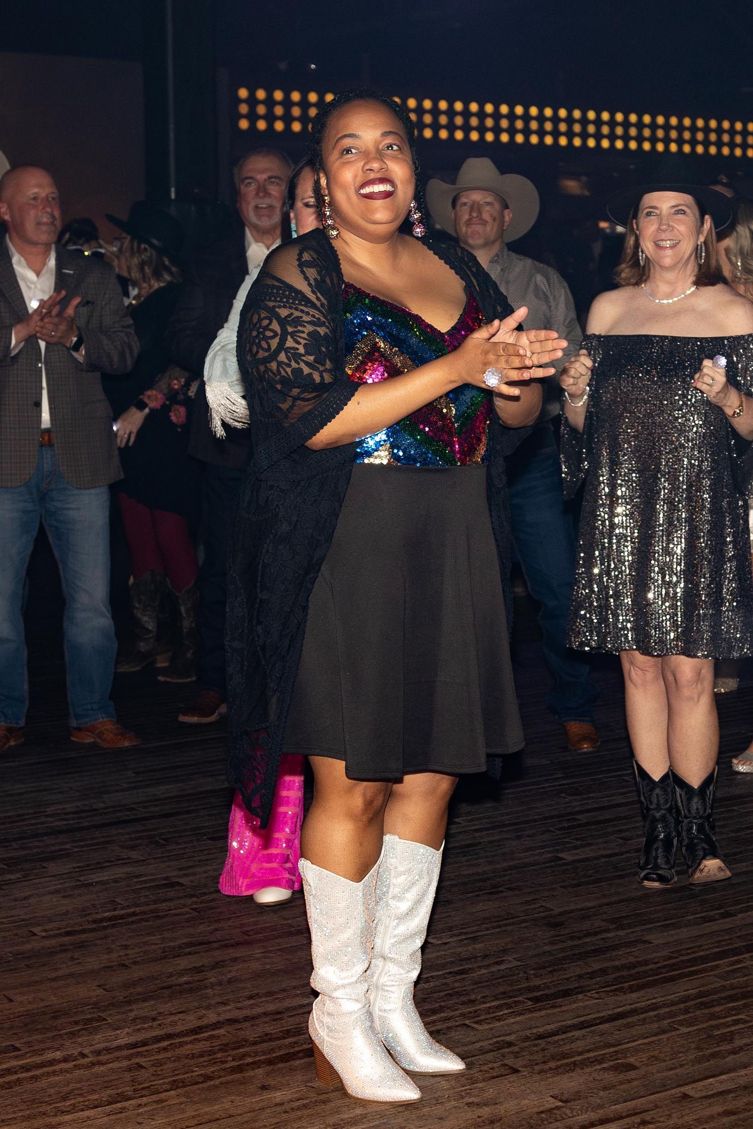 Woman in black dress and white boots clapping at a dance, with other dancers in the background.