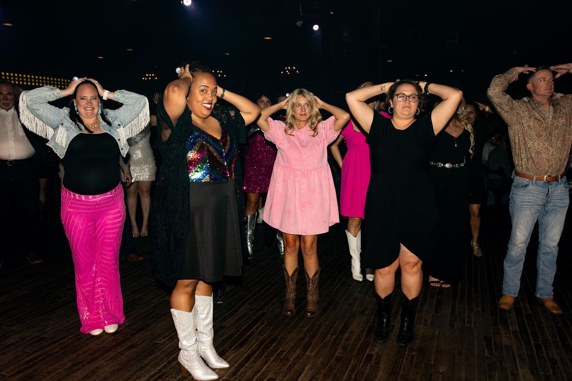 Group of people on a dance floor, arms above their heads, in various outfits.