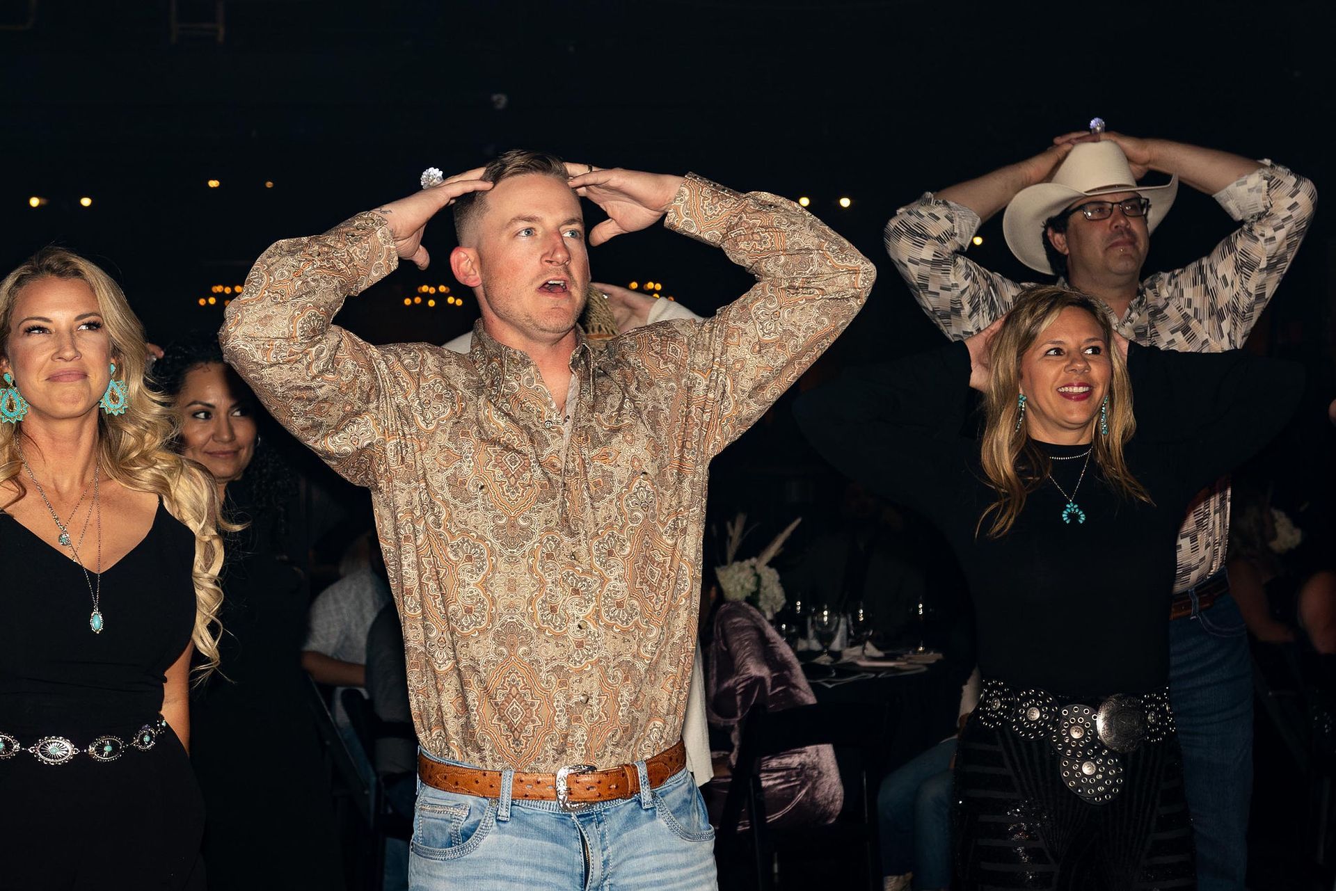People at an event with hands on their heads, one man wearing a patterned shirt, and a woman in a black dress.