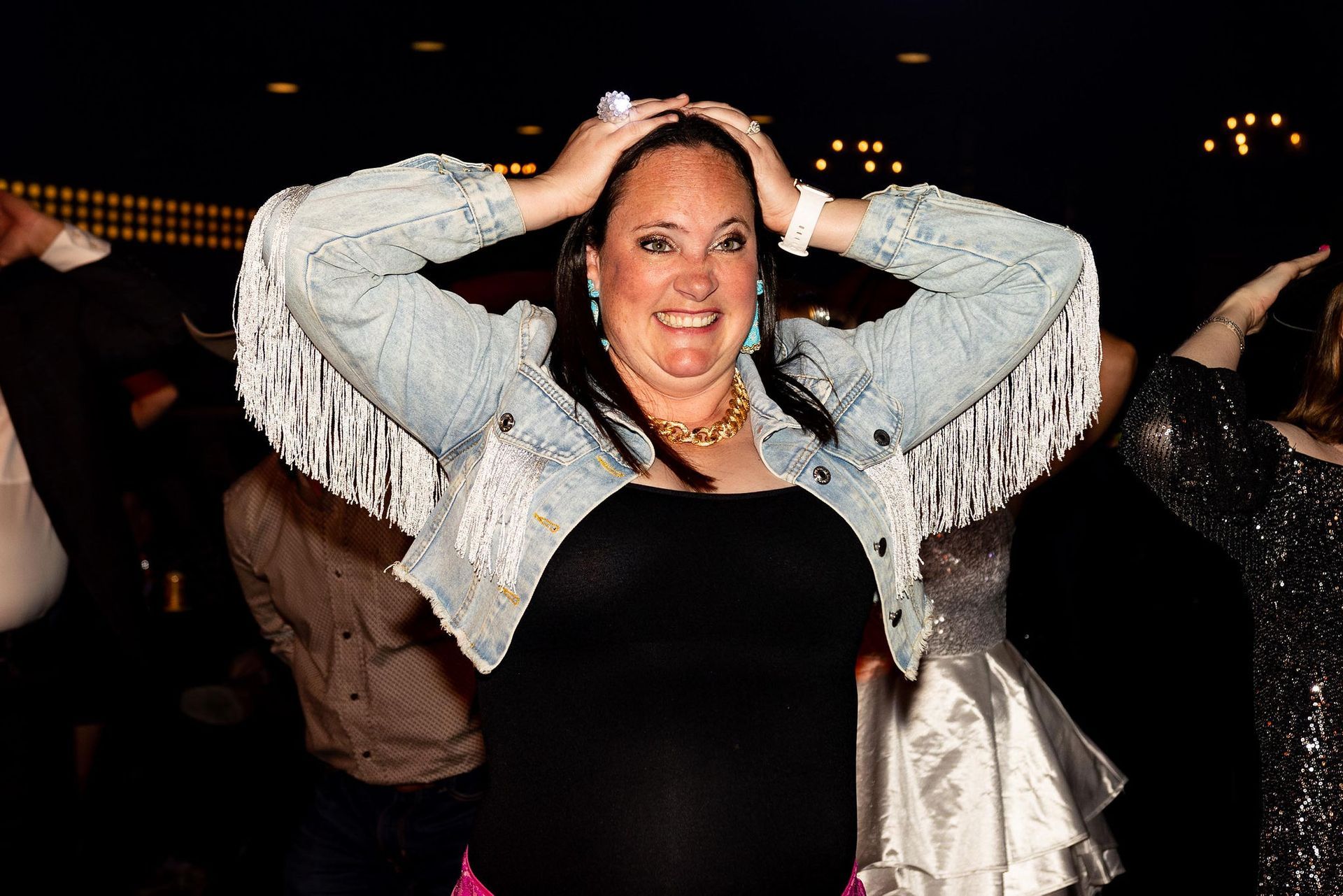 Woman in a fringed denim jacket and black dress, hands on head, smiling in a crowd.