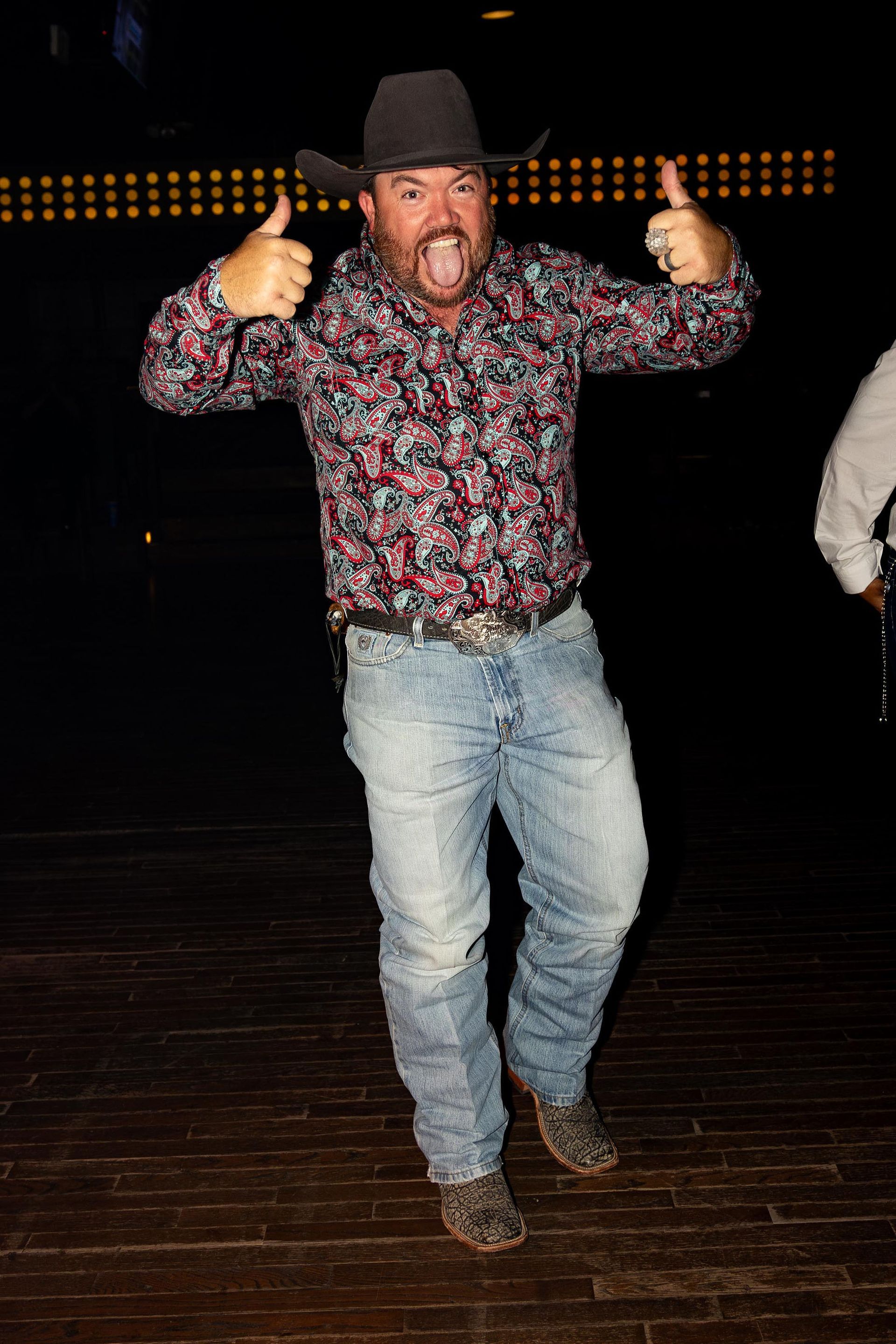 Man in cowboy hat and shirt, giving thumbs up, excited expression. Jeans and boots, dark setting.